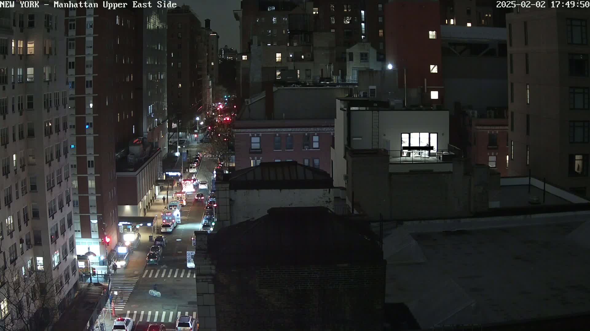 A nighttime, high-angle view of a city street with several buildings and vehicles, some emergency vehicles among them.