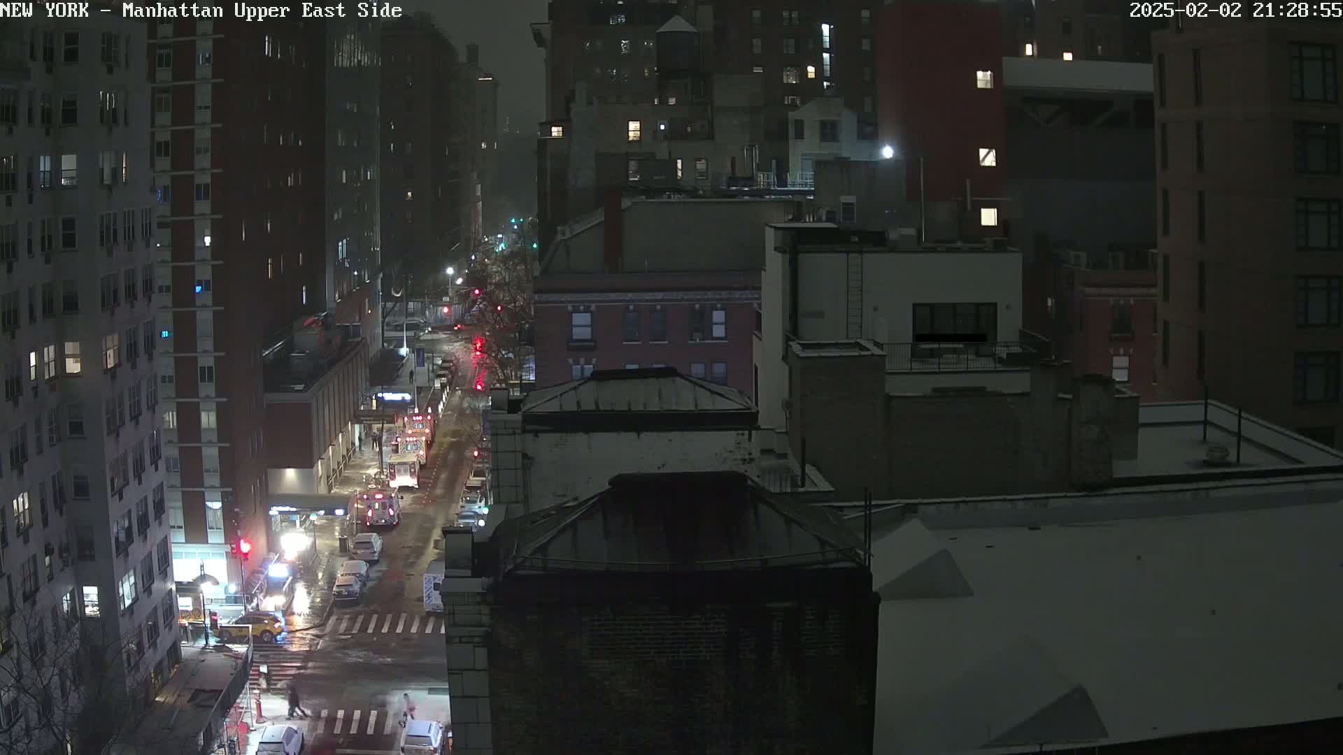 A nighttime, snowy view of city streets and buildings, with emergency vehicles parked on a street below.