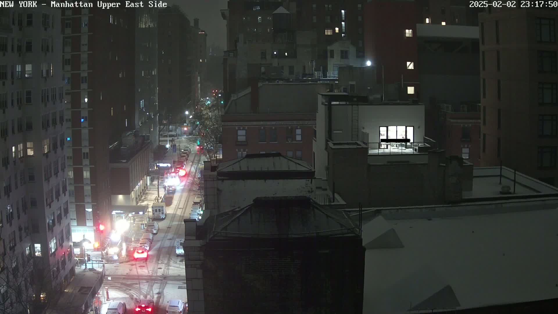 A snow-covered city street at night, viewed from above, shows traffic moving along a street between tall buildings.