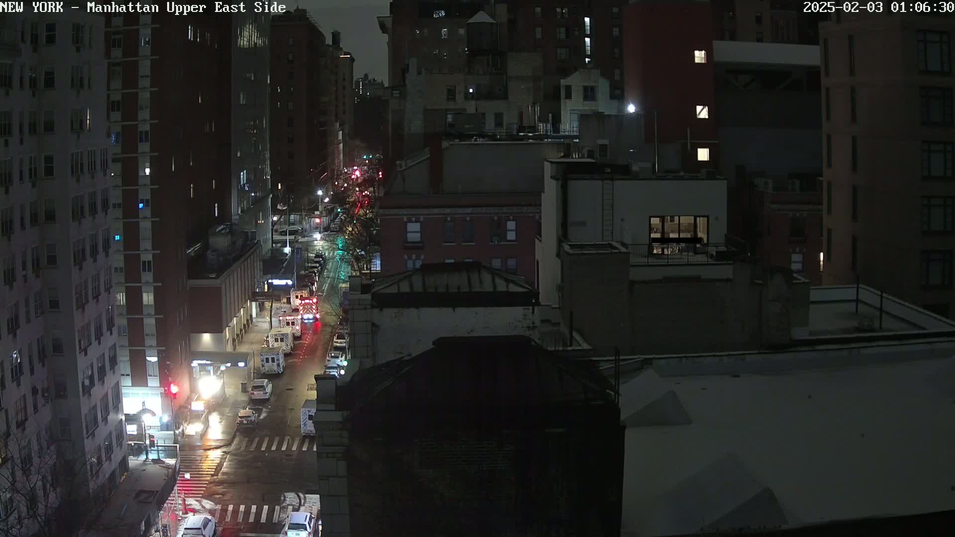 A nighttime aerial view of a city street with several emergency vehicles present, light snow on the ground, and buildings lining the street.