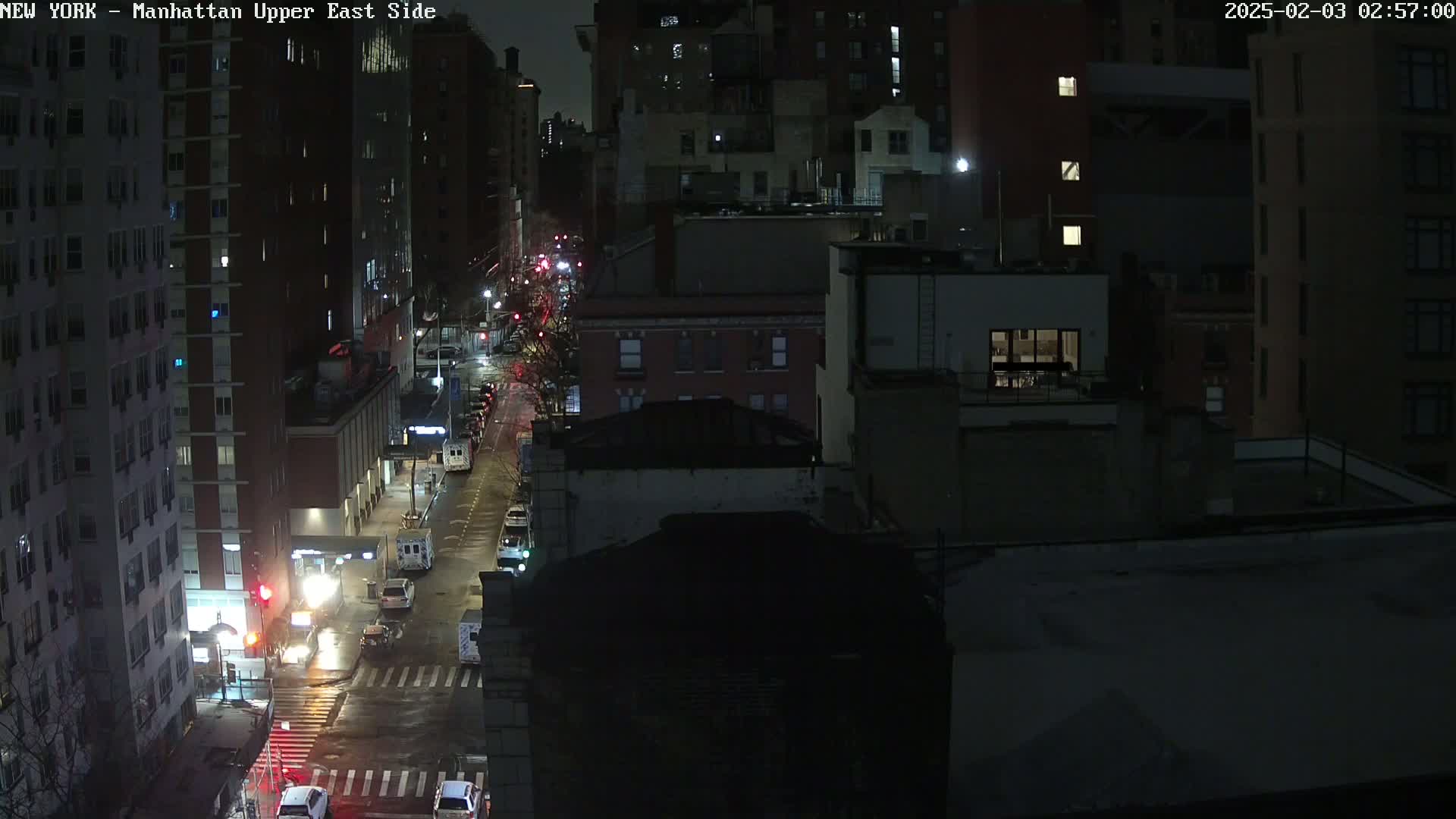 A nighttime, high-angle view of a city street with several cars and buildings, wet from rain.