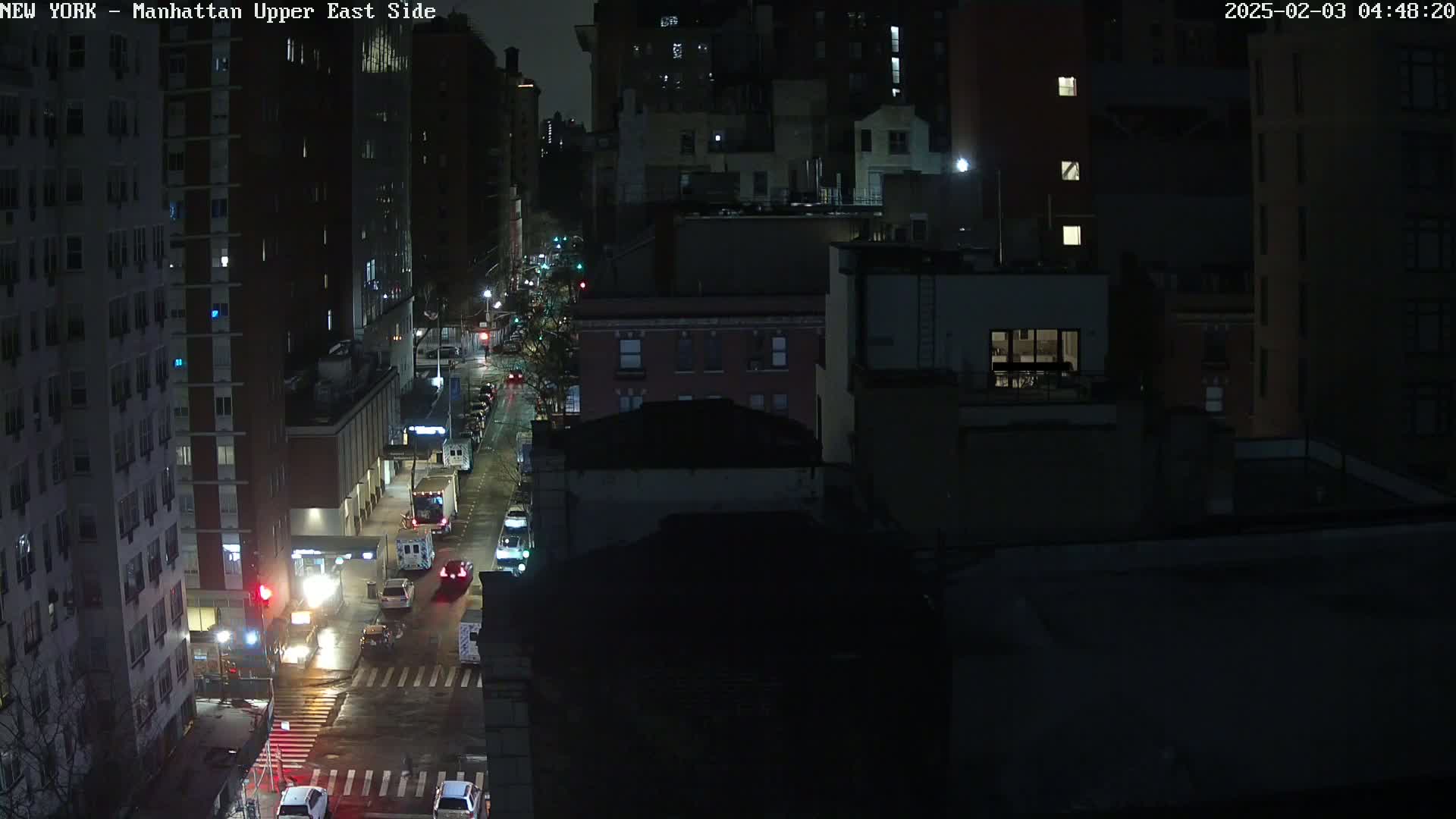 A nighttime, high-angle view of a city street with several cars and buildings, showing wet pavement.