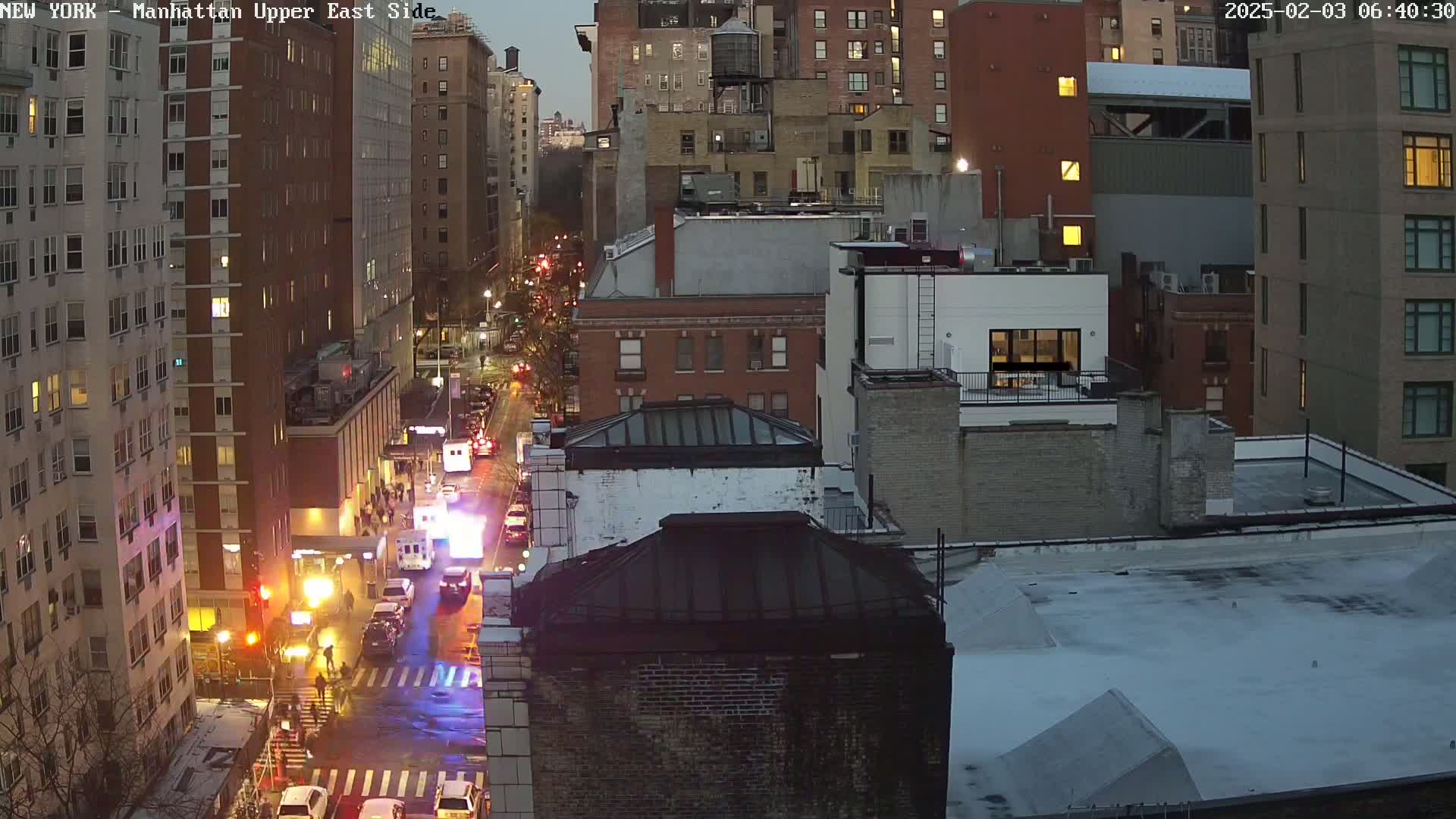 An aerial nighttime view of a city street with emergency vehicles at an intersection, surrounded by tall buildings under a twilight sky.
