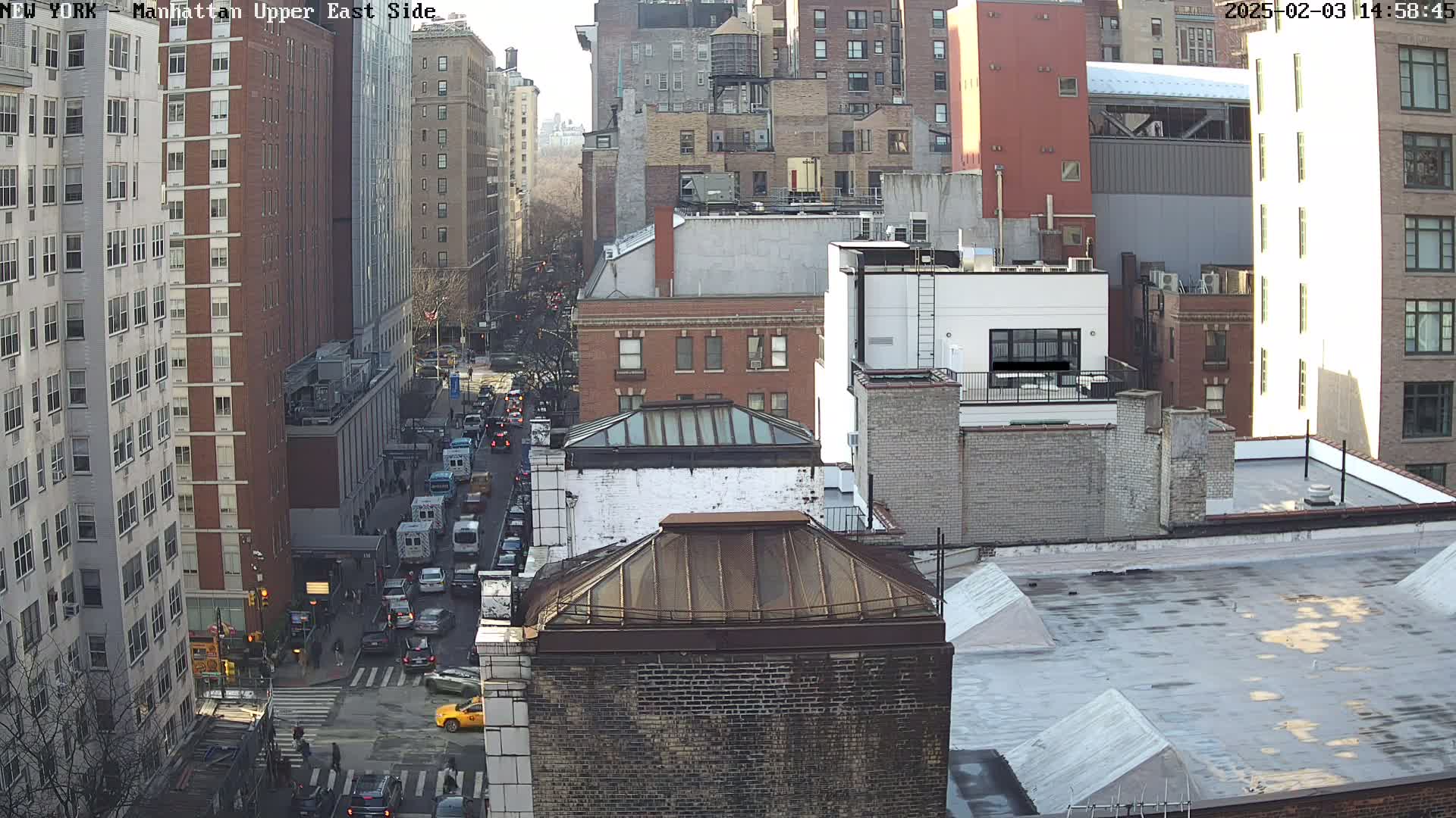 A high-angle view of a city street scene on a sunny day, showing several multi-story buildings and traffic moving along the road below.