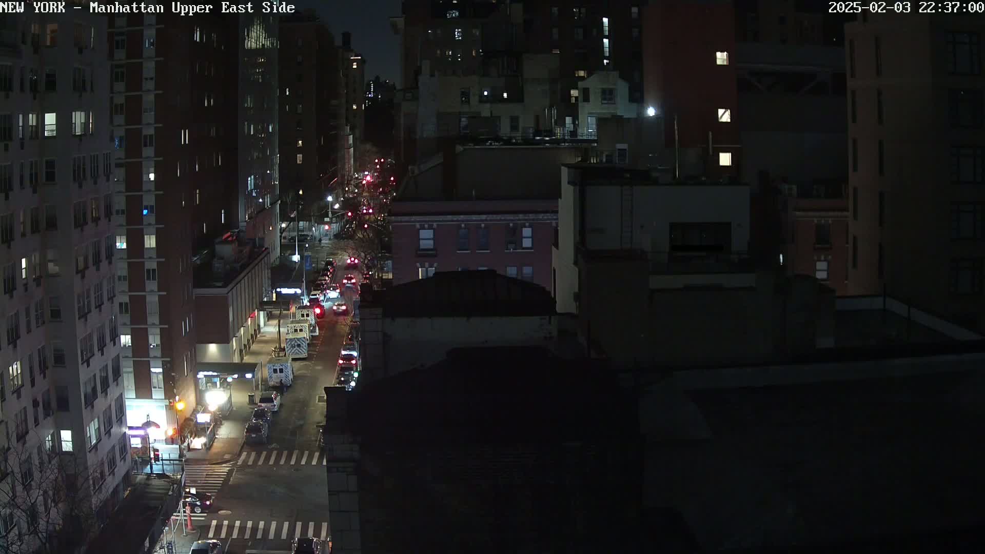 A nighttime aerial view of a city street with several vehicles driving on it, surrounded by tall buildings.