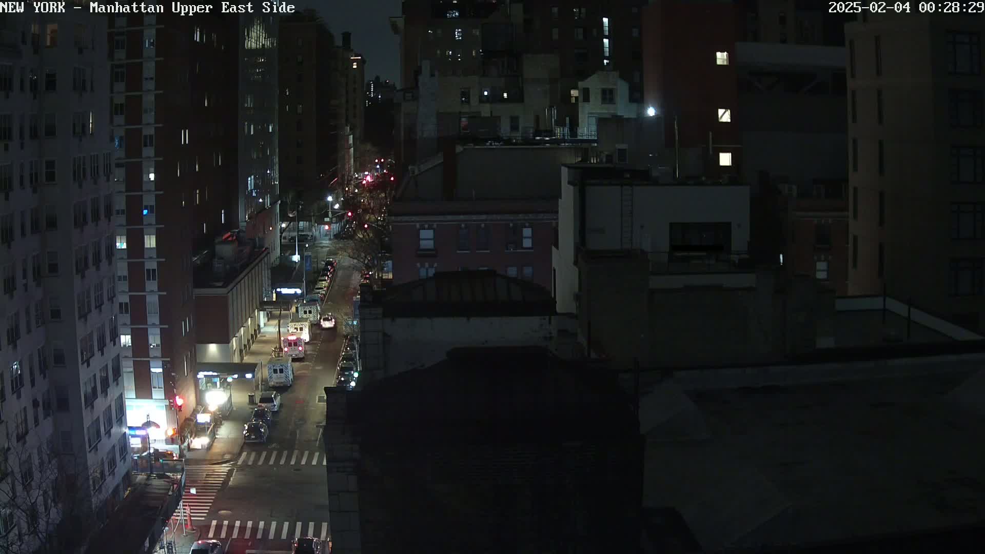 A nighttime aerial view of a city street shows several dark buildings and a few vehicles on a road, under clear skies.