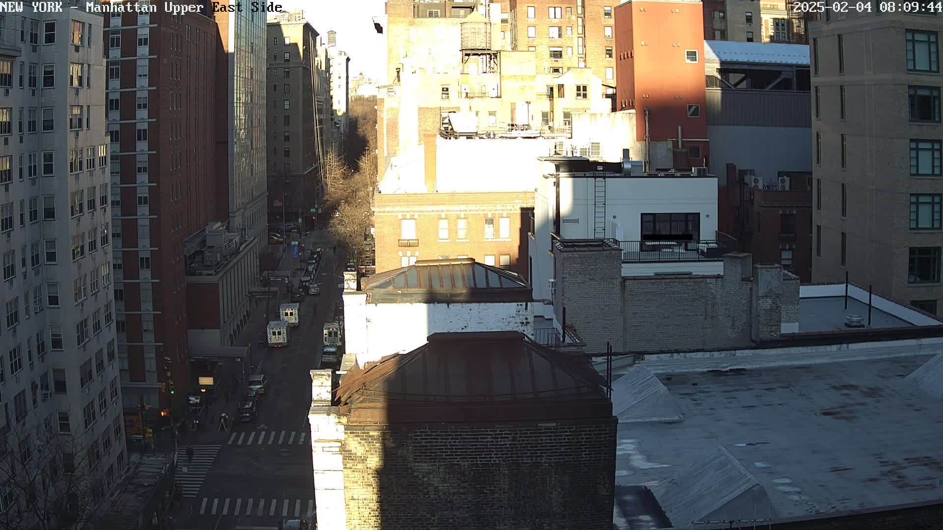 A sunny, high-angle view of a city street lined with tall buildings, showing a mix of architectural styles and some vehicles on the road below.