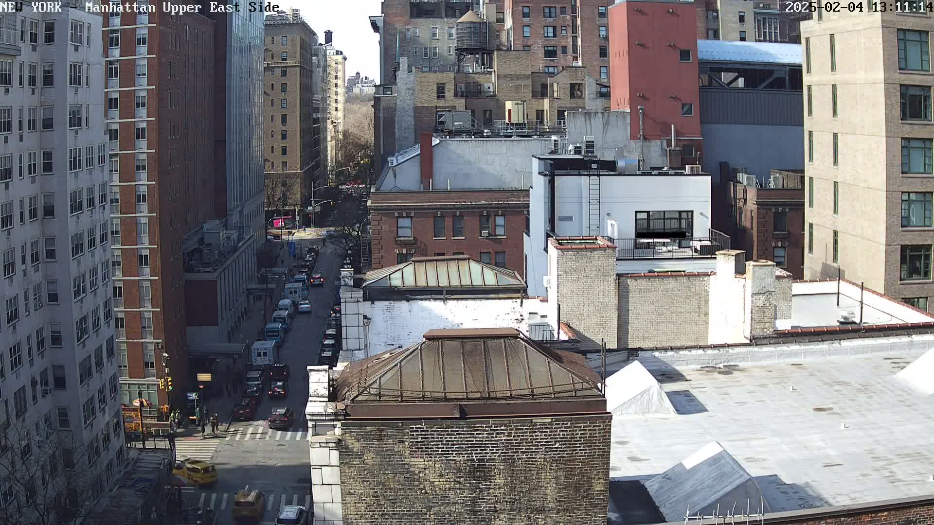 A sunny day reveals a high-angle view of a city street with several multi-story buildings, cars, and pedestrians.