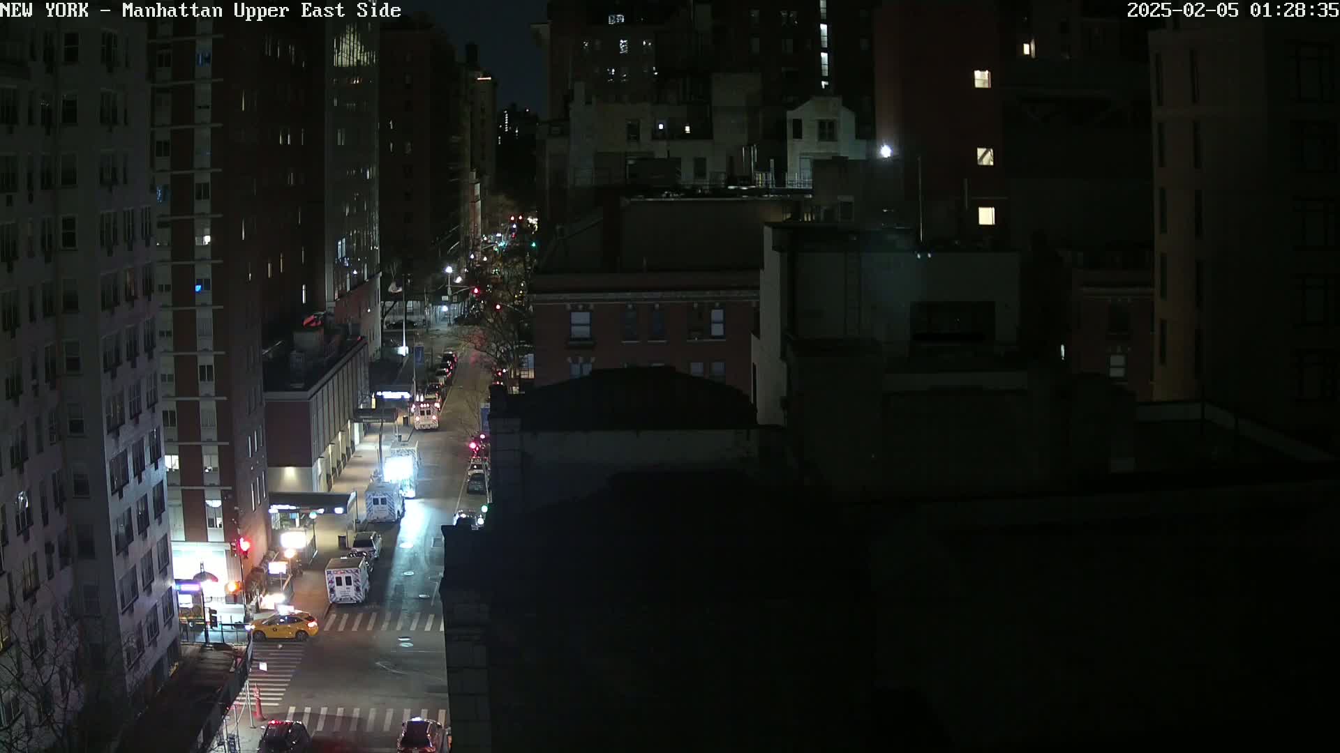 A nighttime, high-angle view of a city street shows several vehicles at an intersection between tall buildings.