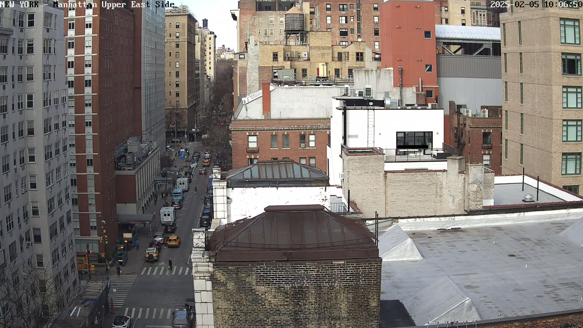A high-angle view of a city street shows several multi-story brick buildings on a partly sunny day with a few cars traveling down the street.