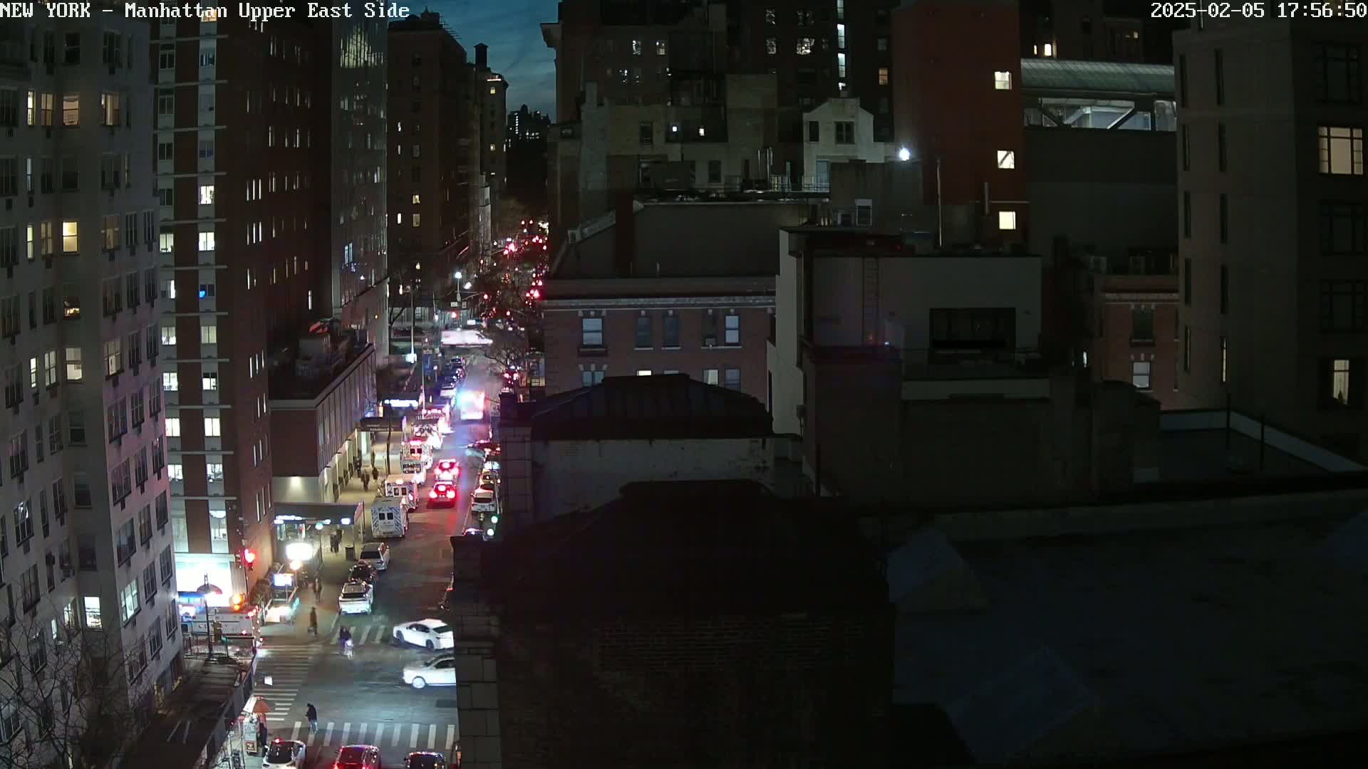 A nighttime, high-angle view of a city street with several emergency vehicles and other cars stopped at an intersection, surrounded by tall buildings.