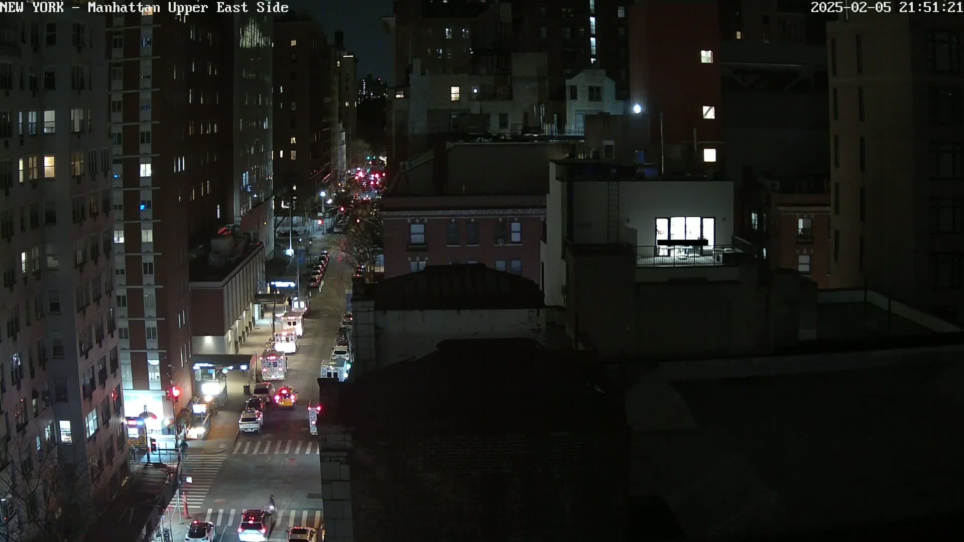 A nighttime aerial view of a city street with several cars and emergency vehicles, showing city buildings lit up against a dark sky.