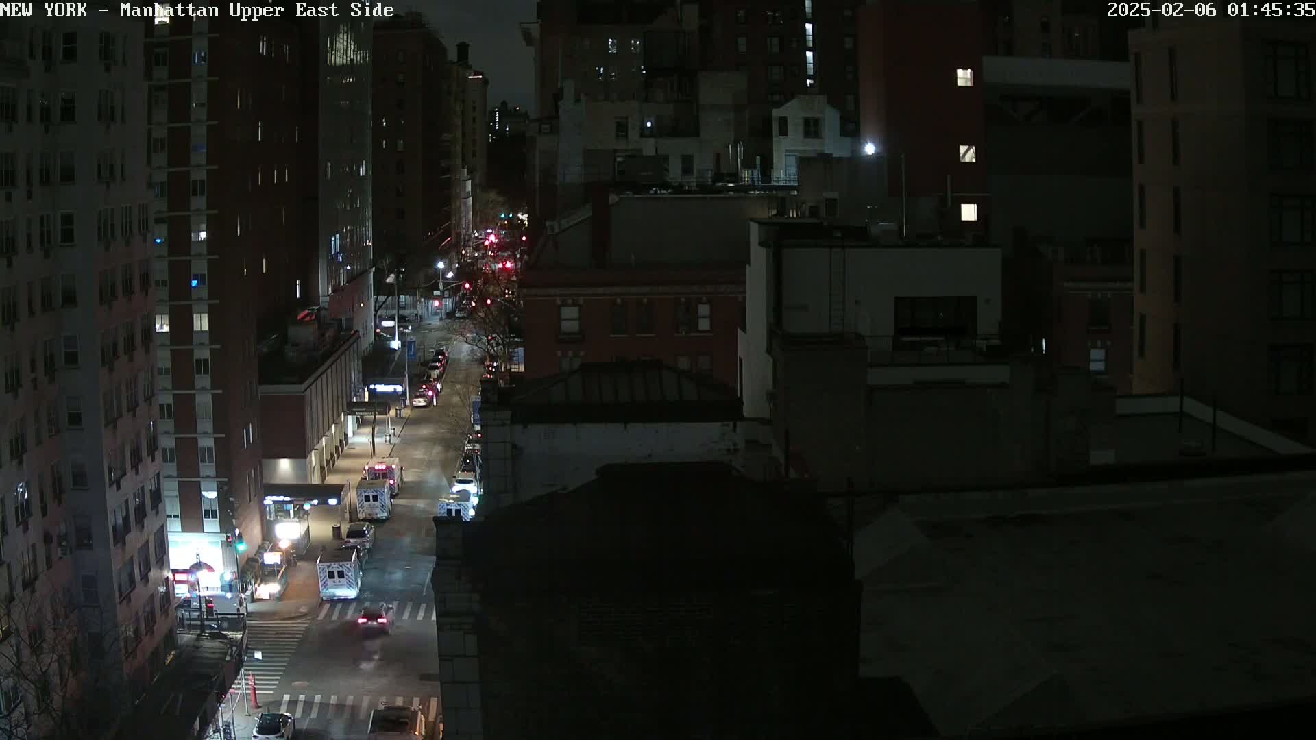 A nighttime, high-angle view of a city street with several vehicles, including what appear to be ambulances, traveling along it, surrounded by dark city buildings.