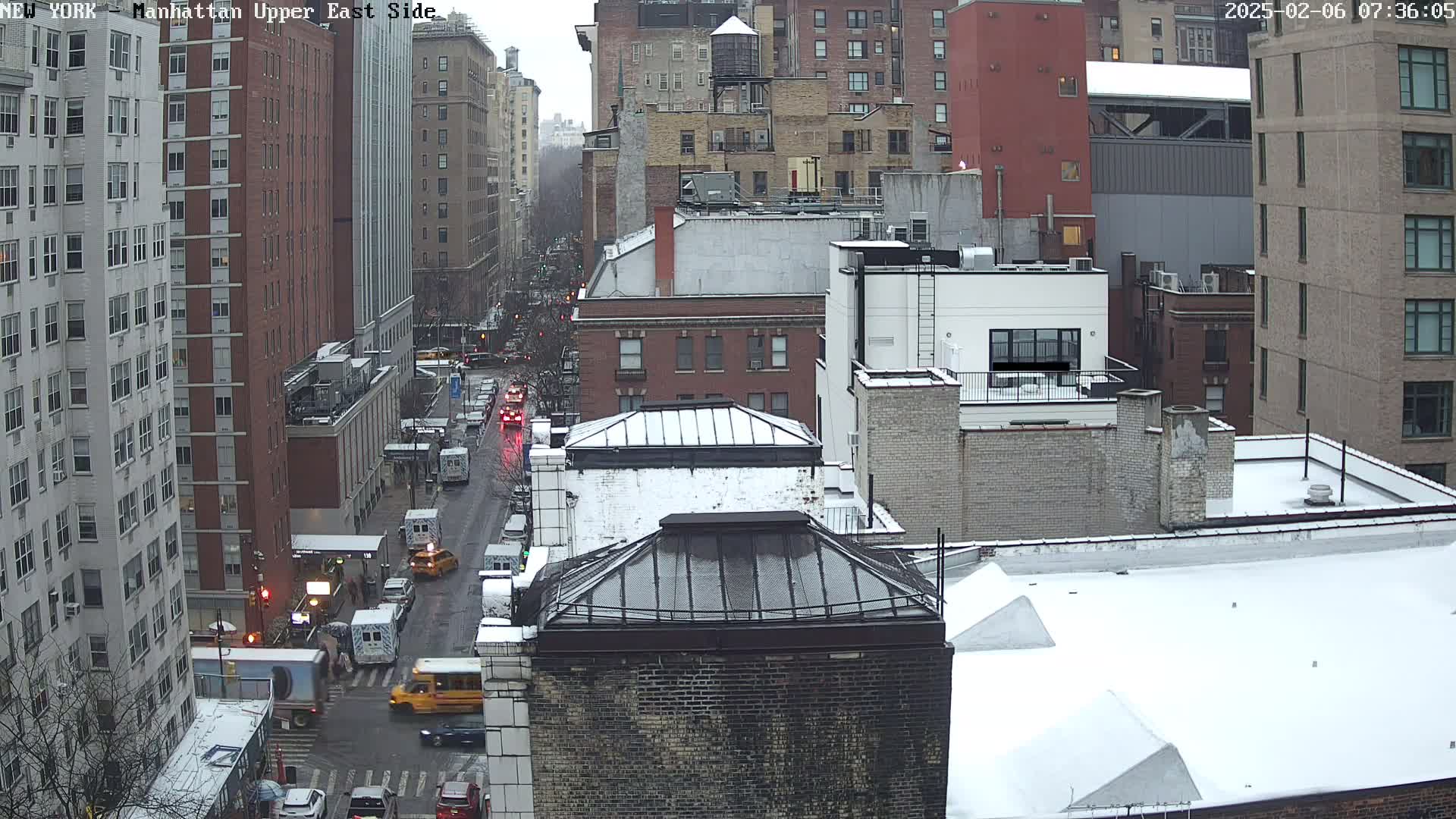 A snowy street scene shows a city block of multi-story buildings, with several vehicles on a street below.
