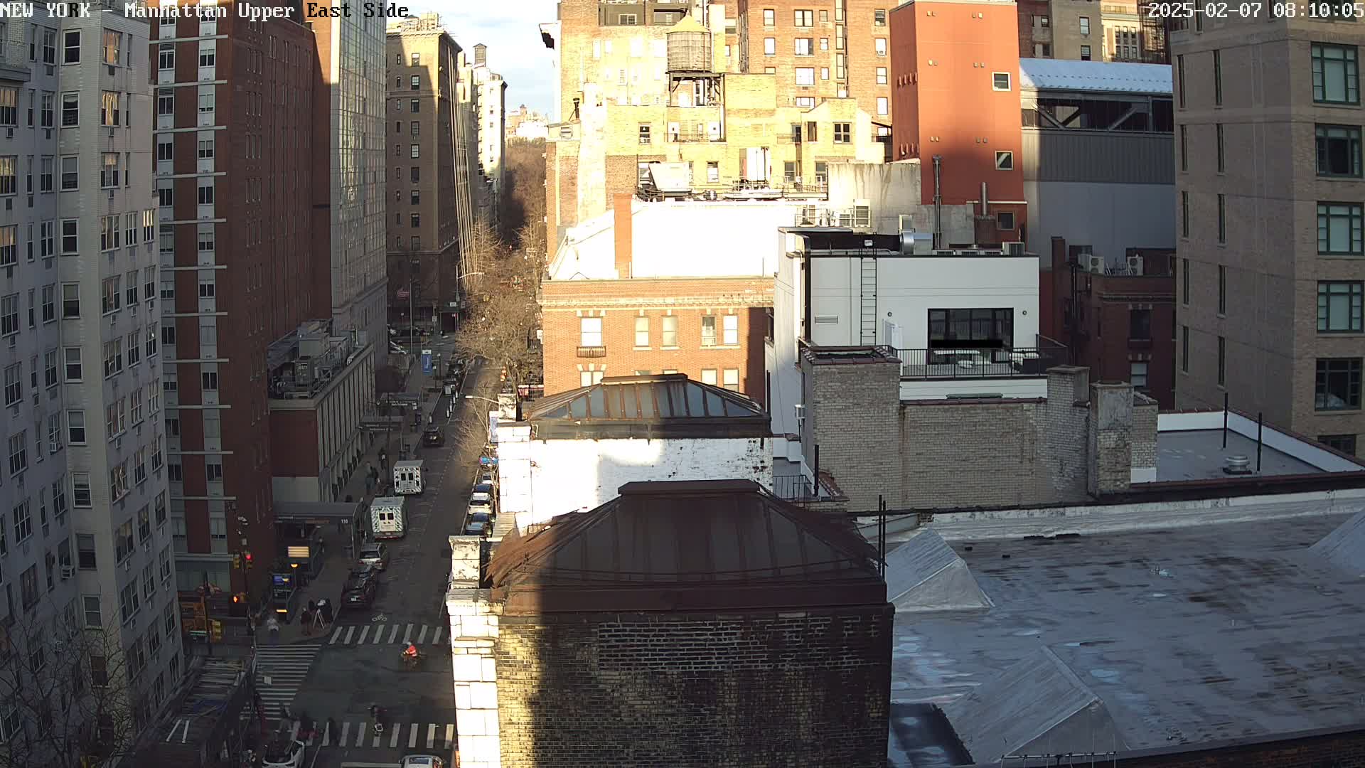 A sunny day view of a city street scene with numerous brick buildings, showing a mostly clear sky and traffic on a street below.