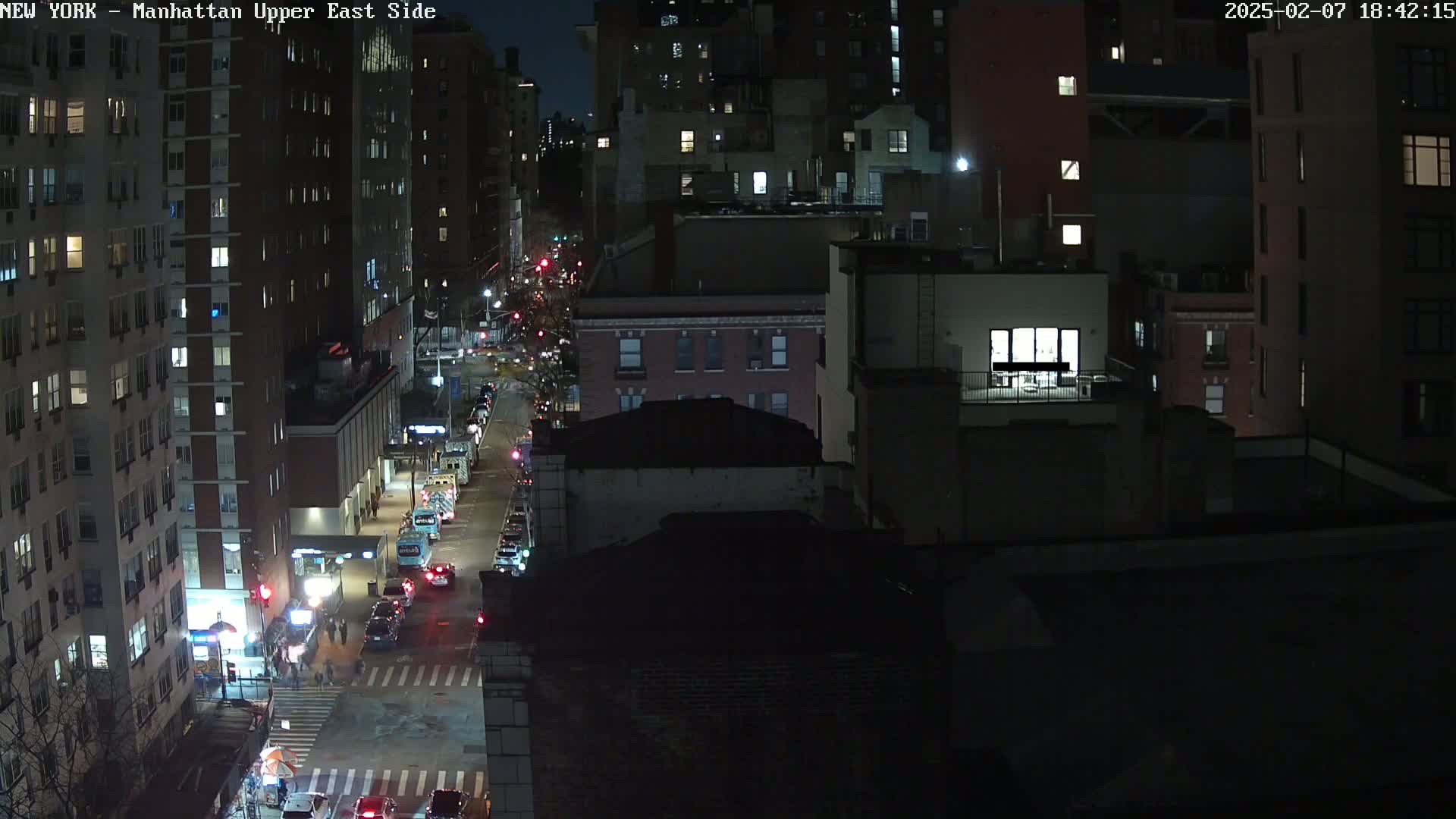 A nighttime aerial view of a city street shows several multi-story buildings with cars driving along a road.
