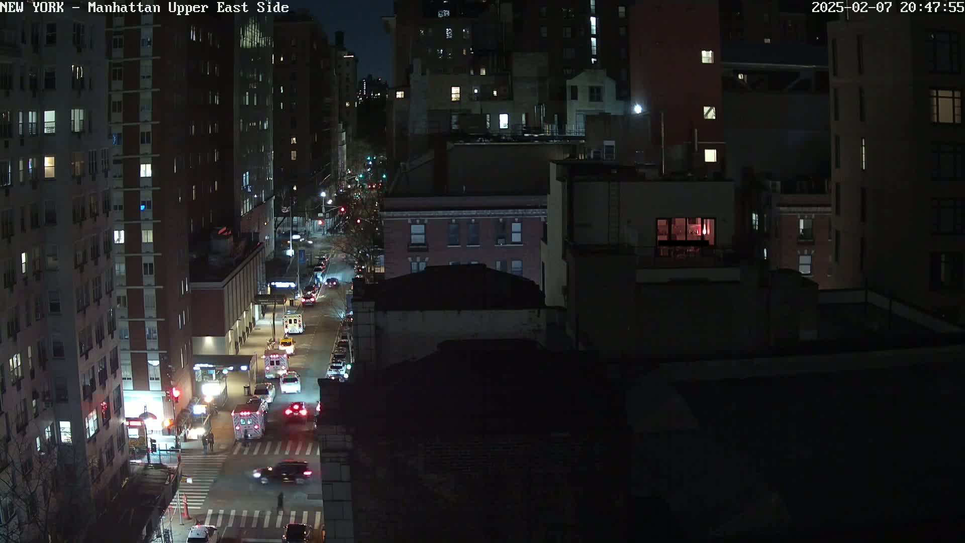 A nighttime aerial view shows a city street with several vehicles, including emergency vehicles, and tall buildings surrounding it under a dark sky.