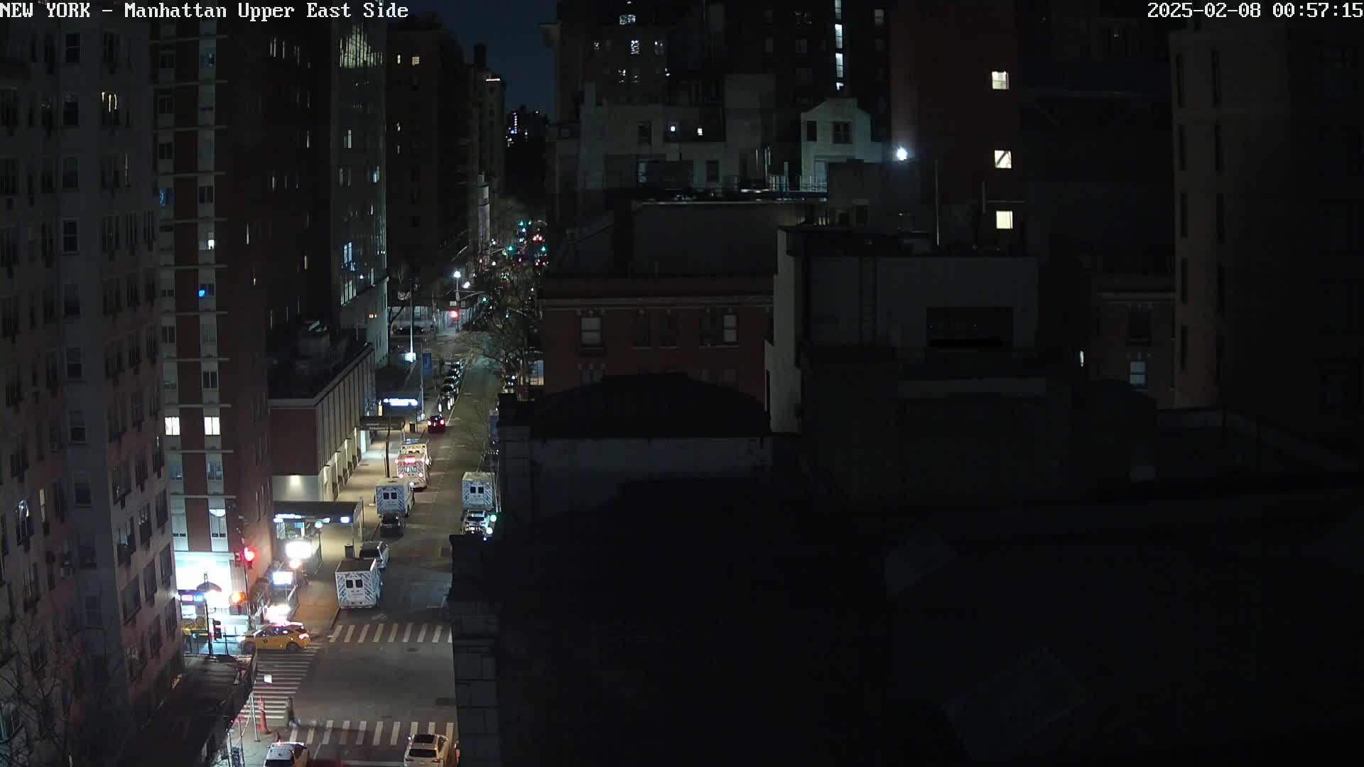 A nighttime aerial view of a city street shows several vehicles, including what appear to be emergency vehicles, stopped at an intersection among dark city buildings under a clear night sky.