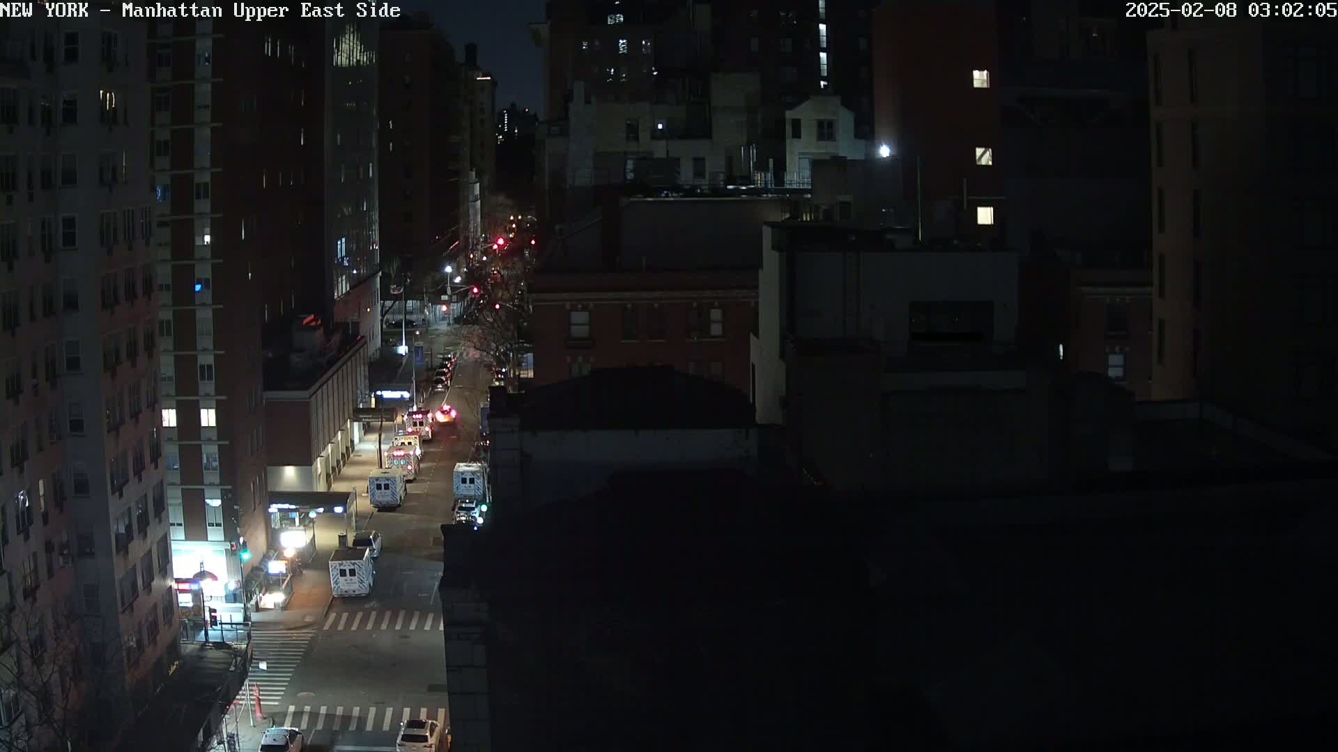 A nighttime, high-angle view shows a city street with several emergency vehicles parked along the side of the road in front of tall buildings.
