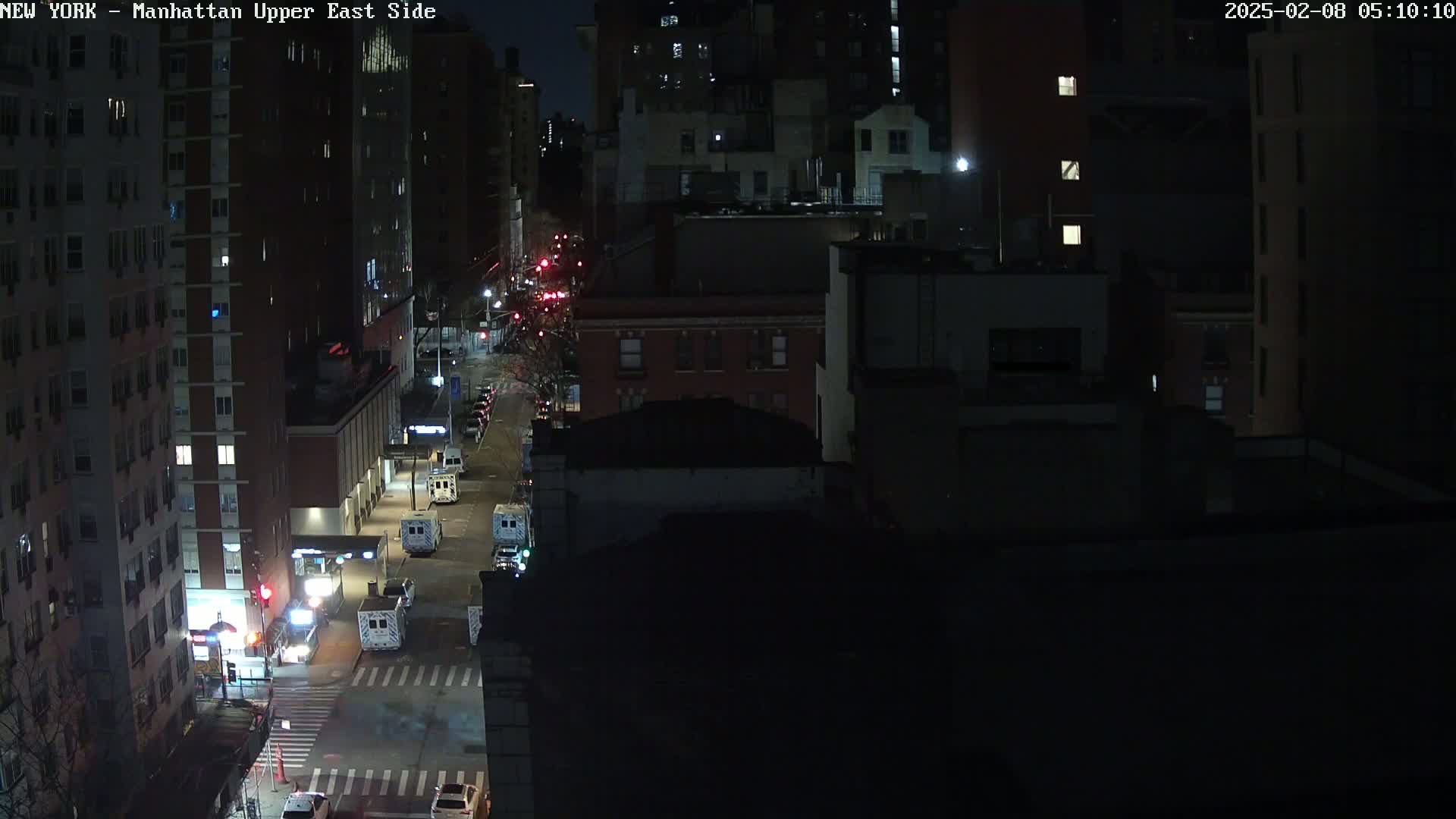 A nighttime aerial view of city streets shows several vehicles parked along the road and a few driving down a multi-lane road, with tall buildings illuminated on either side.