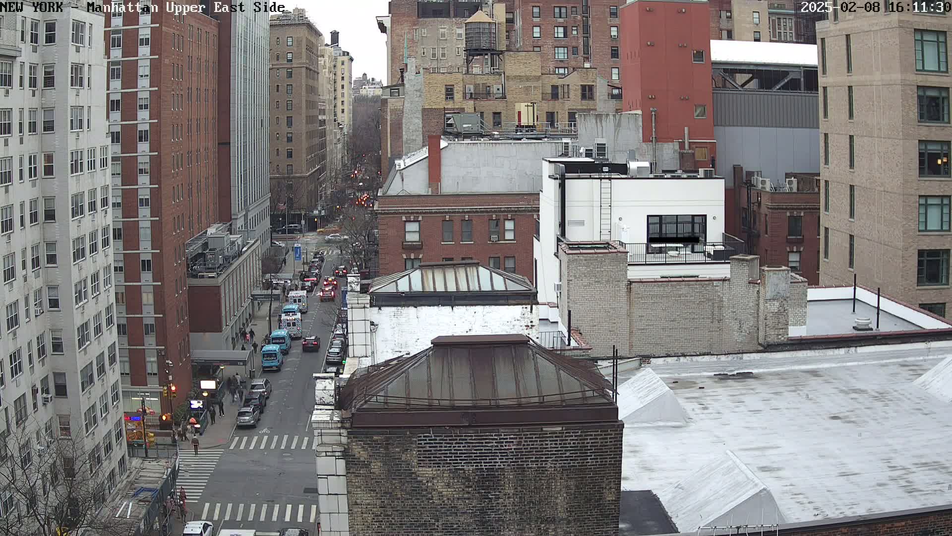 A high-angle view of a city street scene on an overcast day, showing multiple brick buildings, cars parked along the street, and a few pedestrians walking.