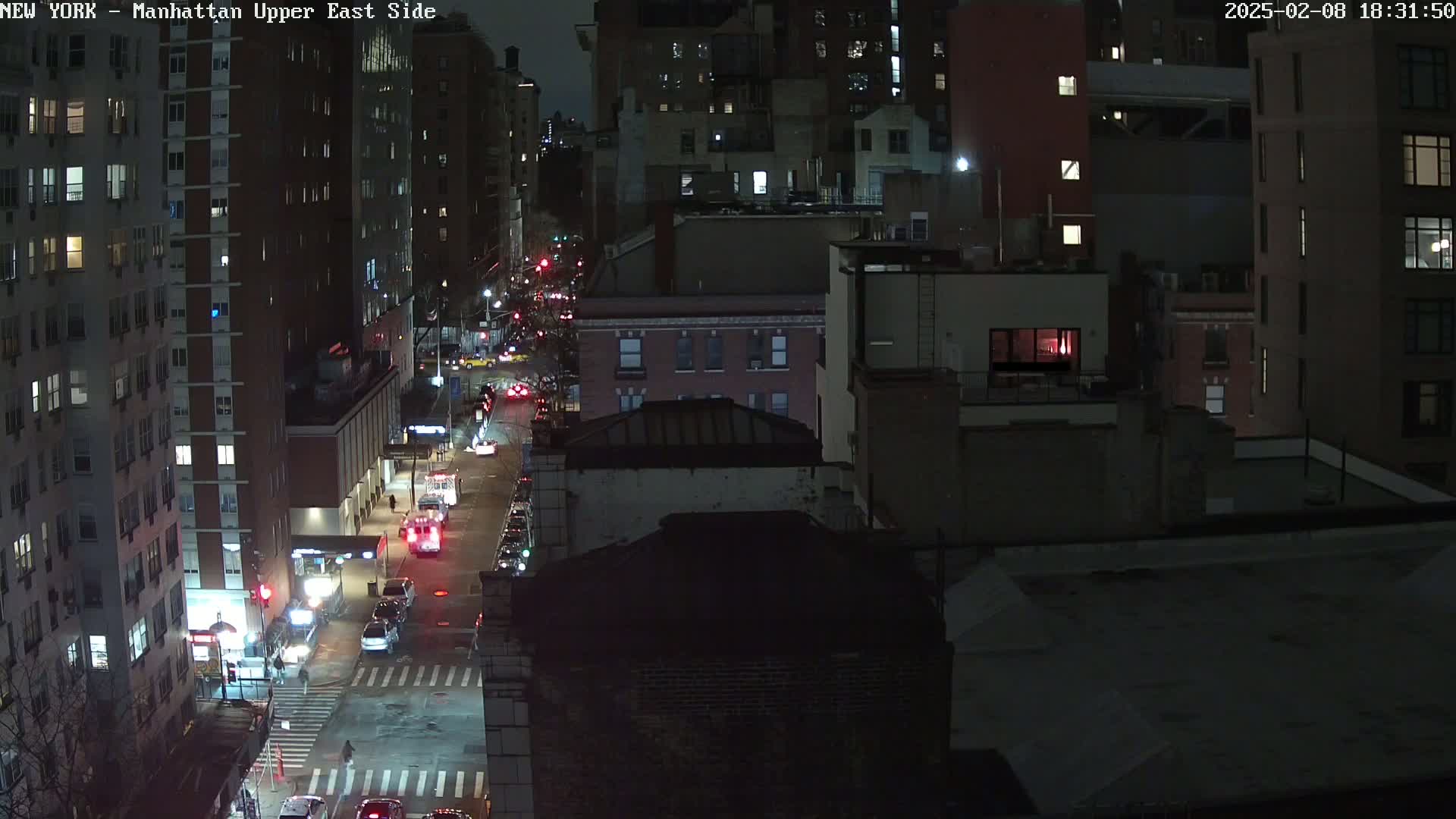 A nighttime aerial view of a city street with vehicles and buildings, under dark conditions.