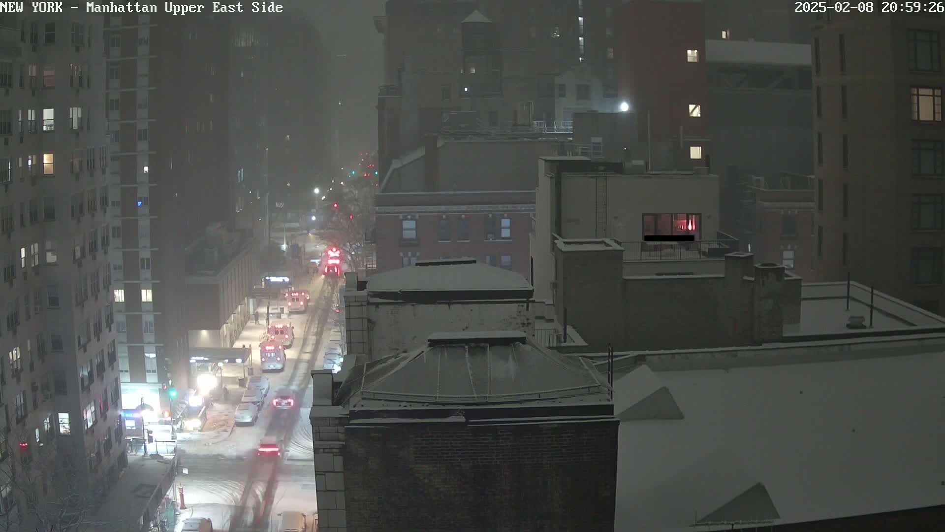 A snow-covered city street at night, with several vehicles, including emergency vehicles, driving through a heavy snowfall.
