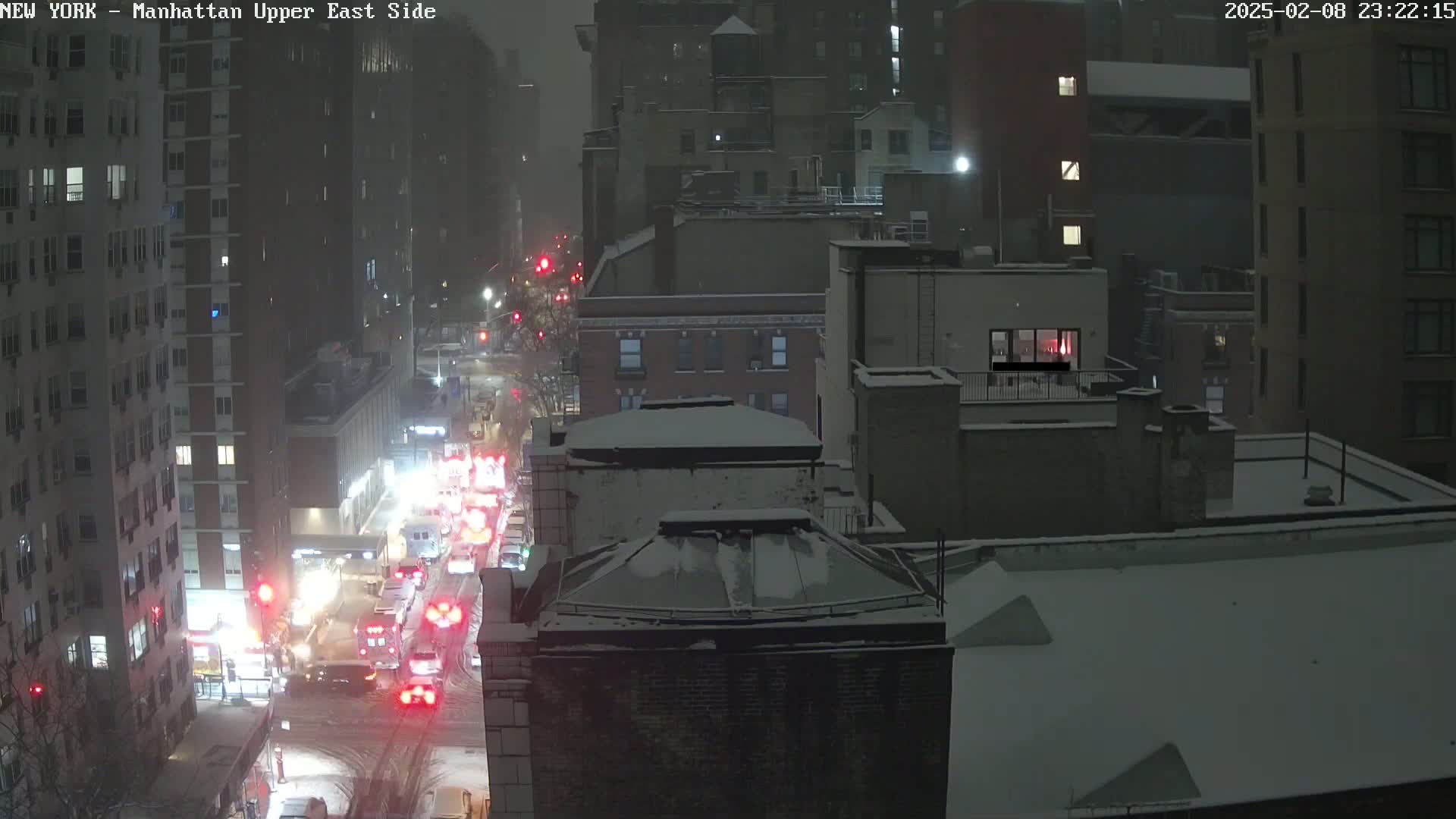 A nighttime, snowy scene shows a city street with traffic, including emergency vehicles, passing between tall buildings covered in snow.