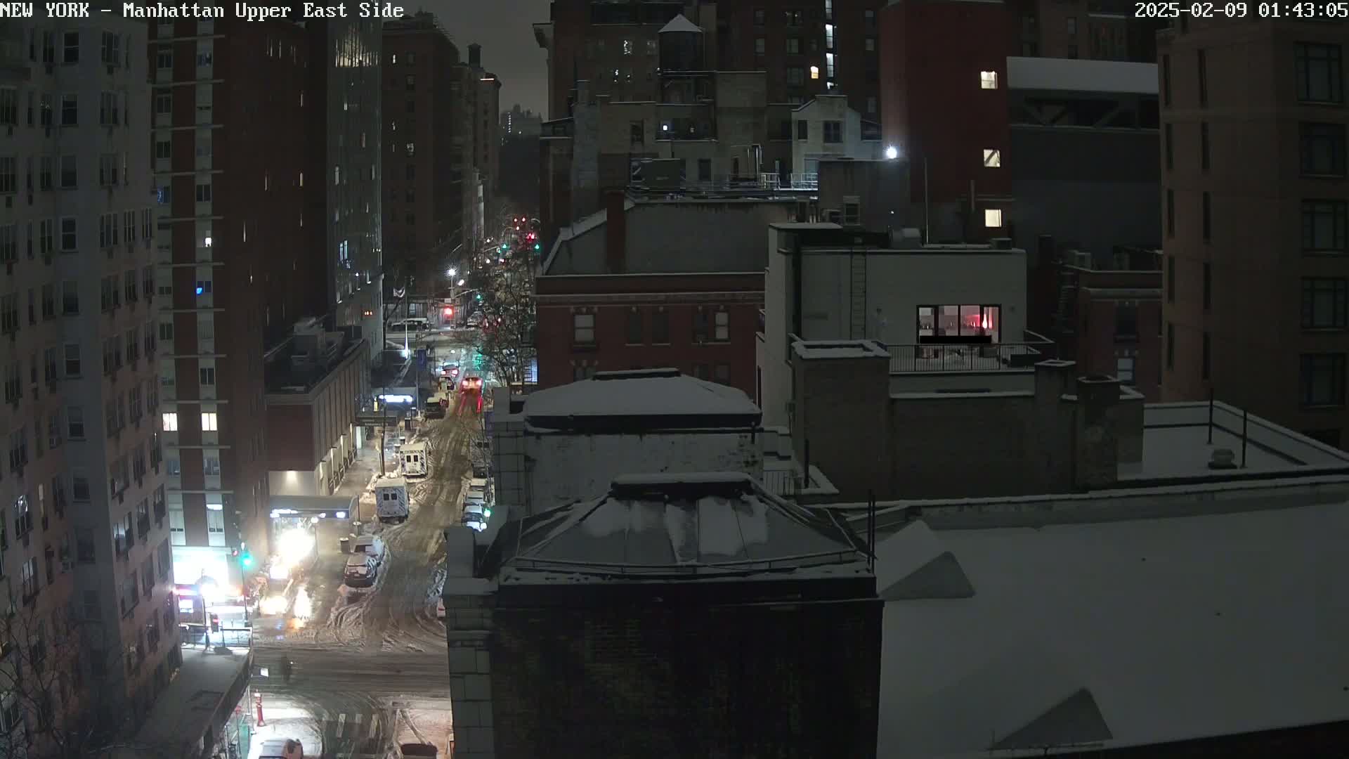 A snow-covered cityscape at night, viewed from above, shows a street with sparsely moving vehicles and apartment buildings with illuminated windows.