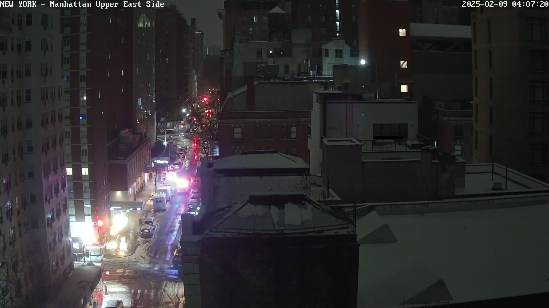 A nighttime, snowy scene shows a city street with emergency vehicles and traffic, surrounded by snow-covered buildings.