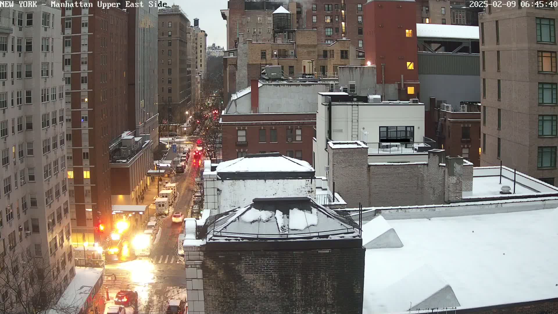 A snowy city street scene showing several multi-story buildings with cars driving on a wet road.