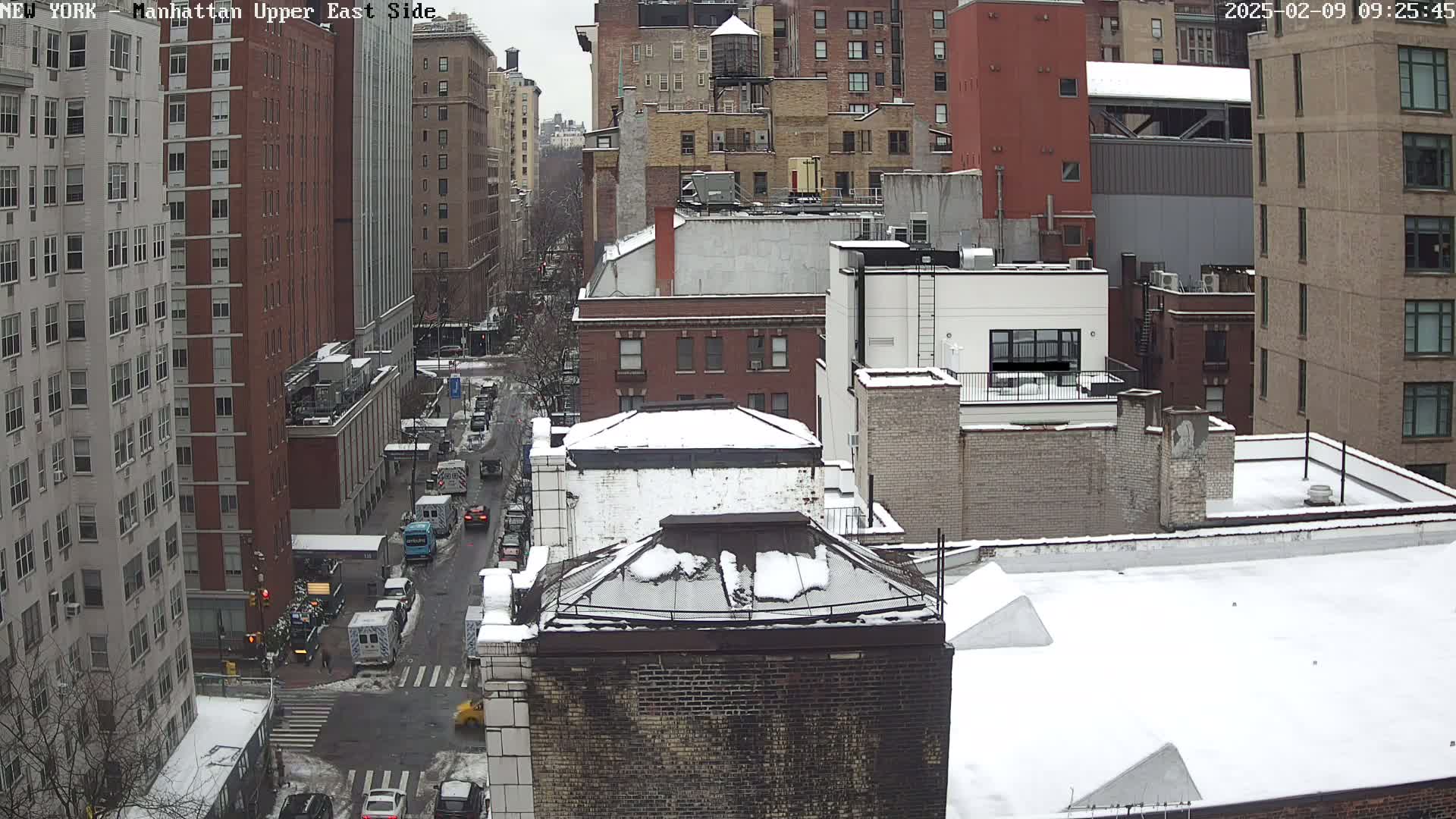 A snow-covered cityscape view of several multi-story brick buildings, with a street below showing sparse traffic.