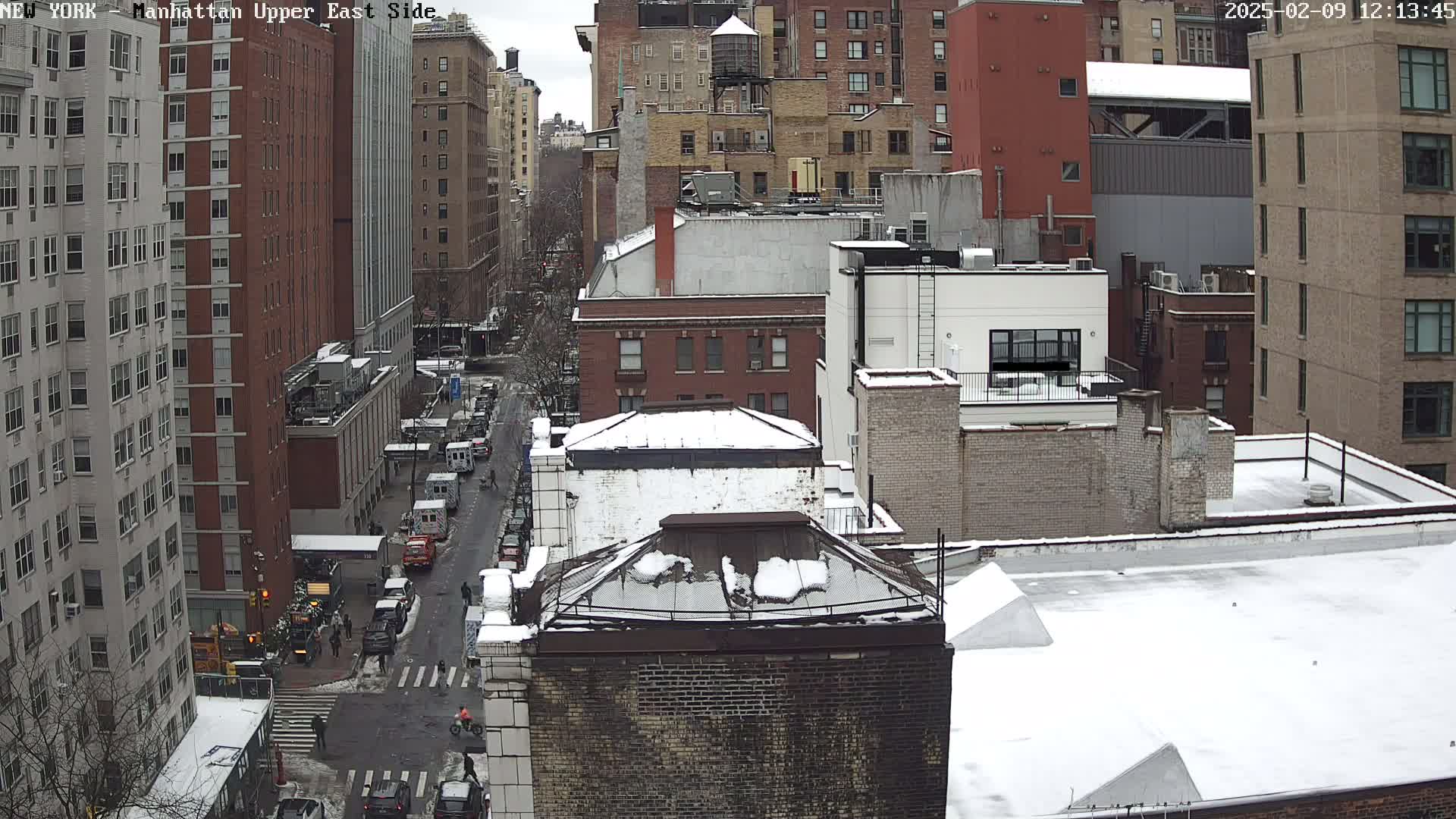 A snow-covered cityscape view shows a street intersection with several cars and pedestrians, alongside multi-story brick buildings.
