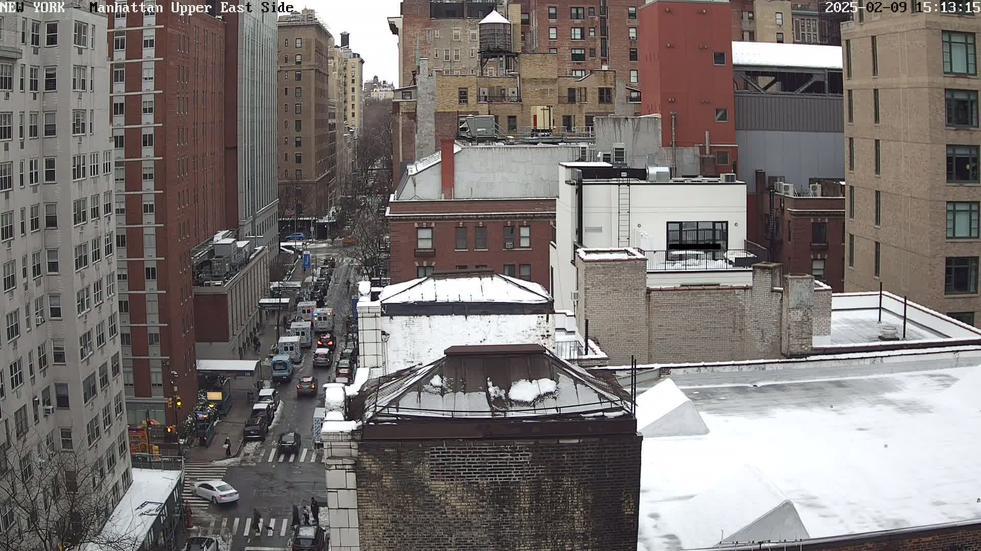 A snow-covered cityscape view shows a street with cars and pedestrians, and numerous brick buildings of varying heights.
