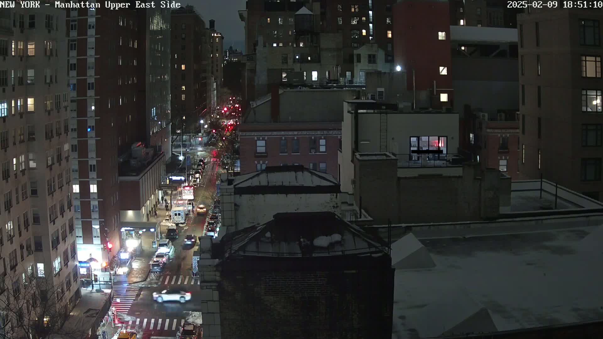 A nighttime, snowy view of city streets and buildings with cars driving on a wet road.