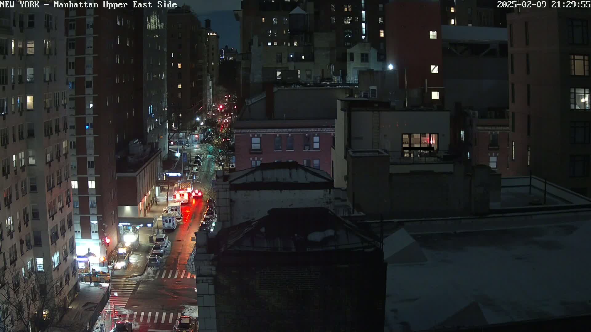 A nighttime aerial view of a city street with several buildings and emergency vehicles, lightly snow-covered.