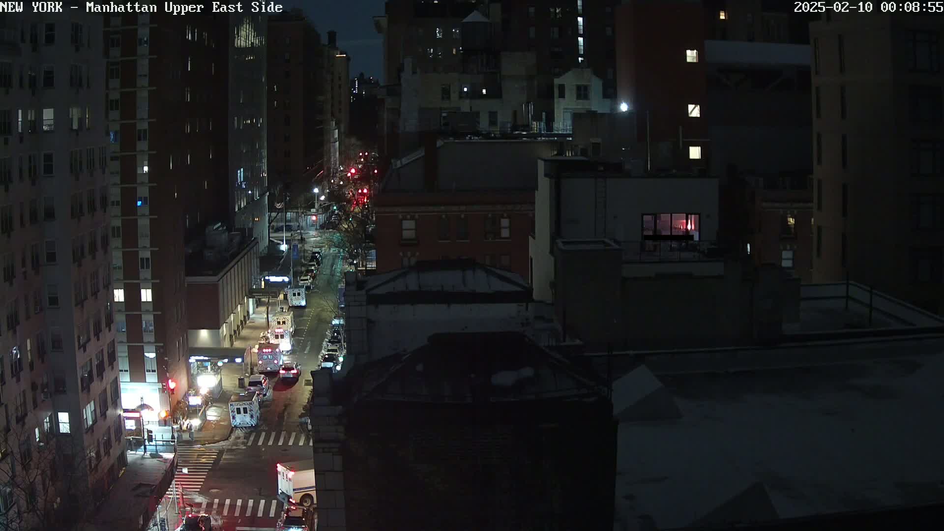A nighttime, snow-dusted city street scene shows several emergency vehicles parked along a roadway between tall buildings.
