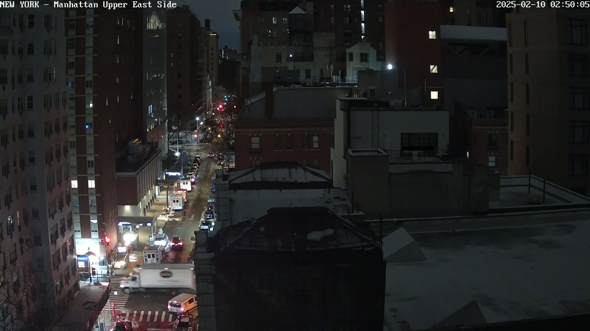 A nighttime, snow-dusted city street scene shows a lightly trafficked road lined with tall buildings, some with visible lights.