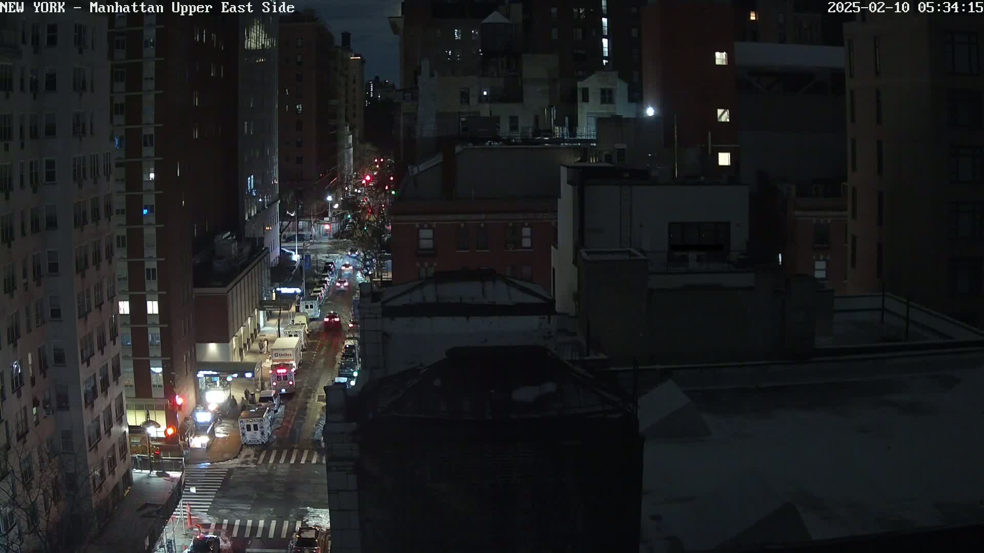 A nighttime aerial view of a city street with sparsely trafficked vehicles, illuminated buildings, and snow-covered ground.