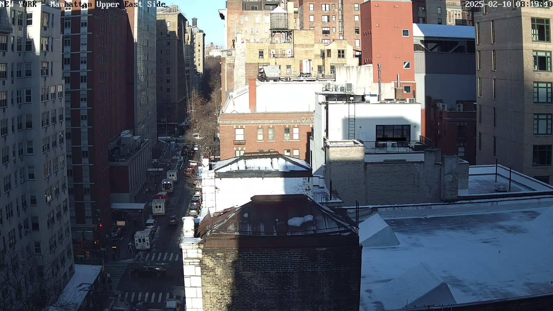 A sunny day with snow on the rooftops of several city buildings, and emergency vehicles are parked on a street below.