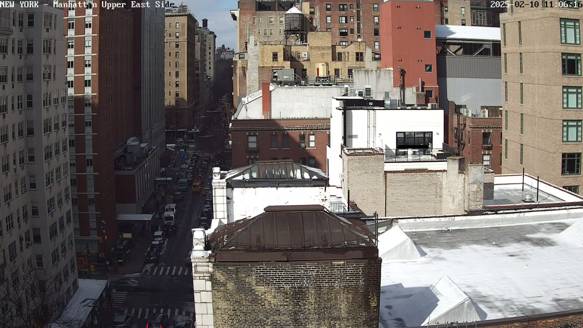 A high-angle view of a city street scene shows numerous brick buildings, some with snow-covered roofs, under a sunny sky.