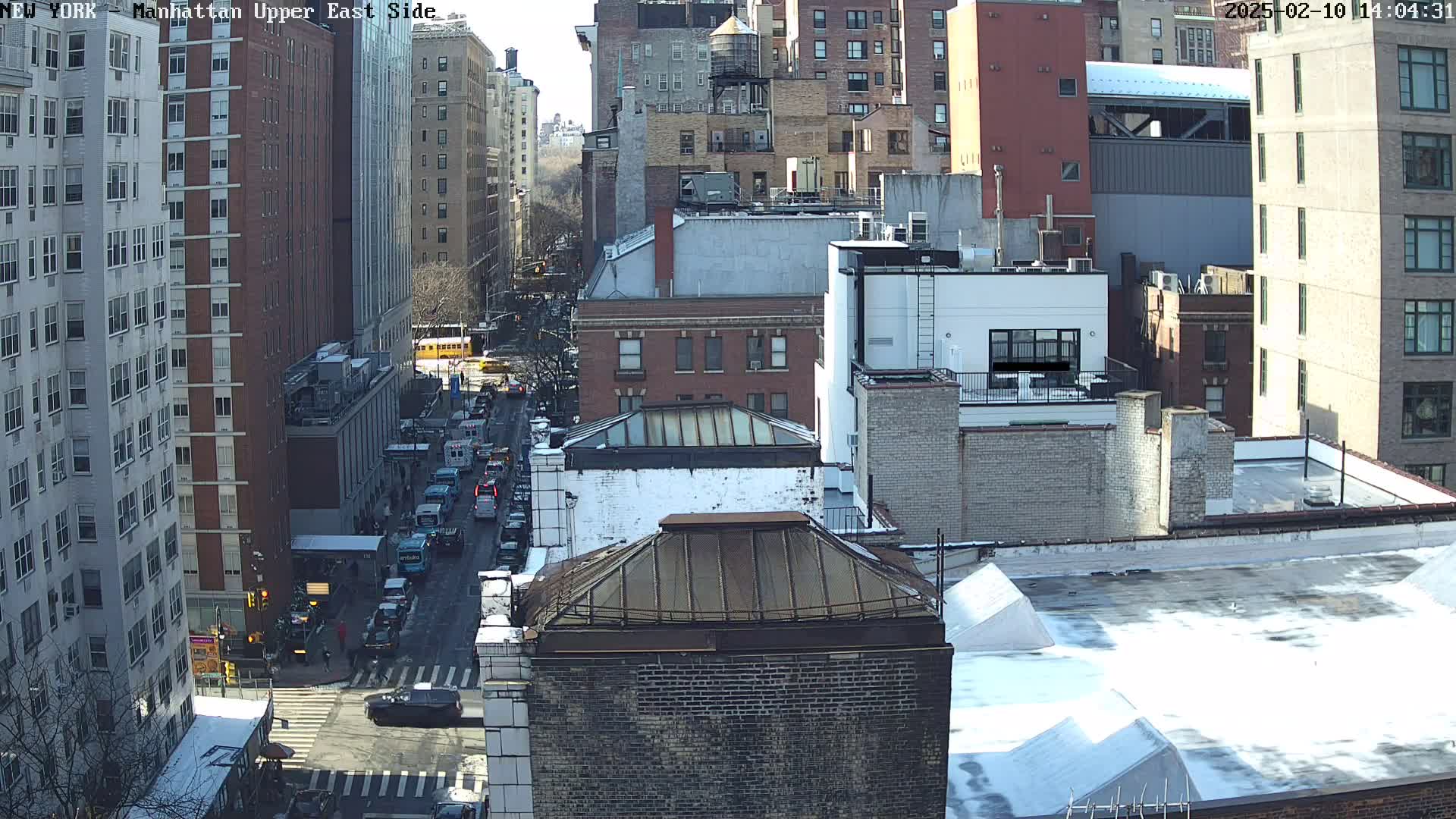 A sunny, winter day view of a city street, showing numerous multi-story buildings, some with snow on their roofs, and vehicles on a street below.