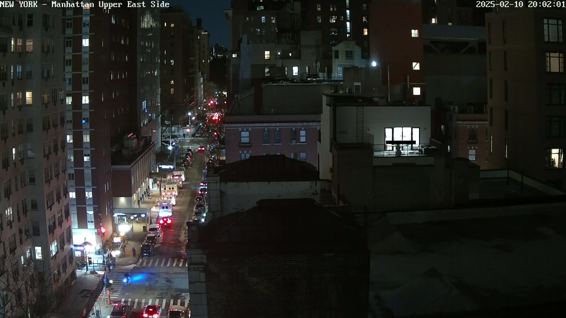 A nighttime aerial view of city streets with cars driving on a road between tall buildings under a dark sky.