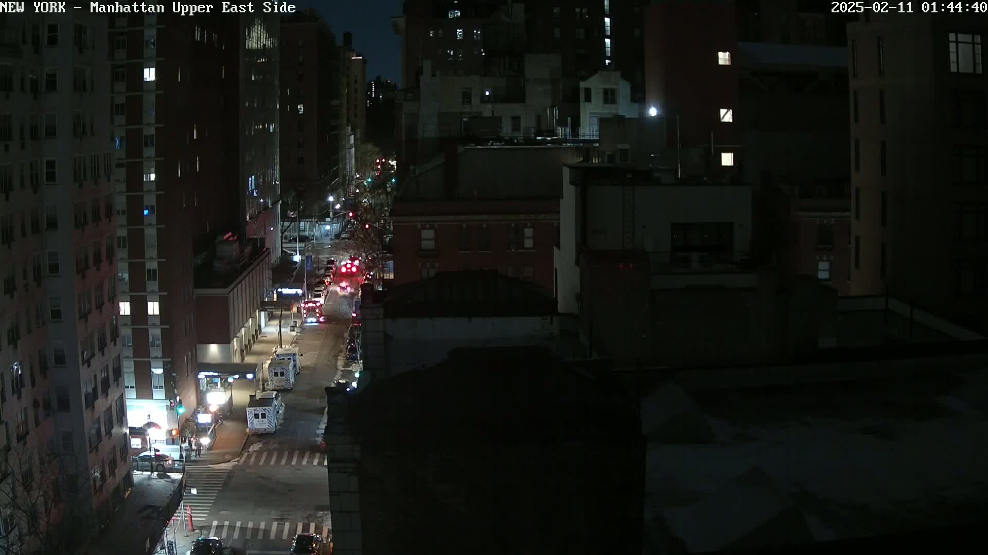A nighttime, high-angle view of a city street with several vehicles, including emergency vehicles, on a snow-dusted road, bordered by multi-story buildings.