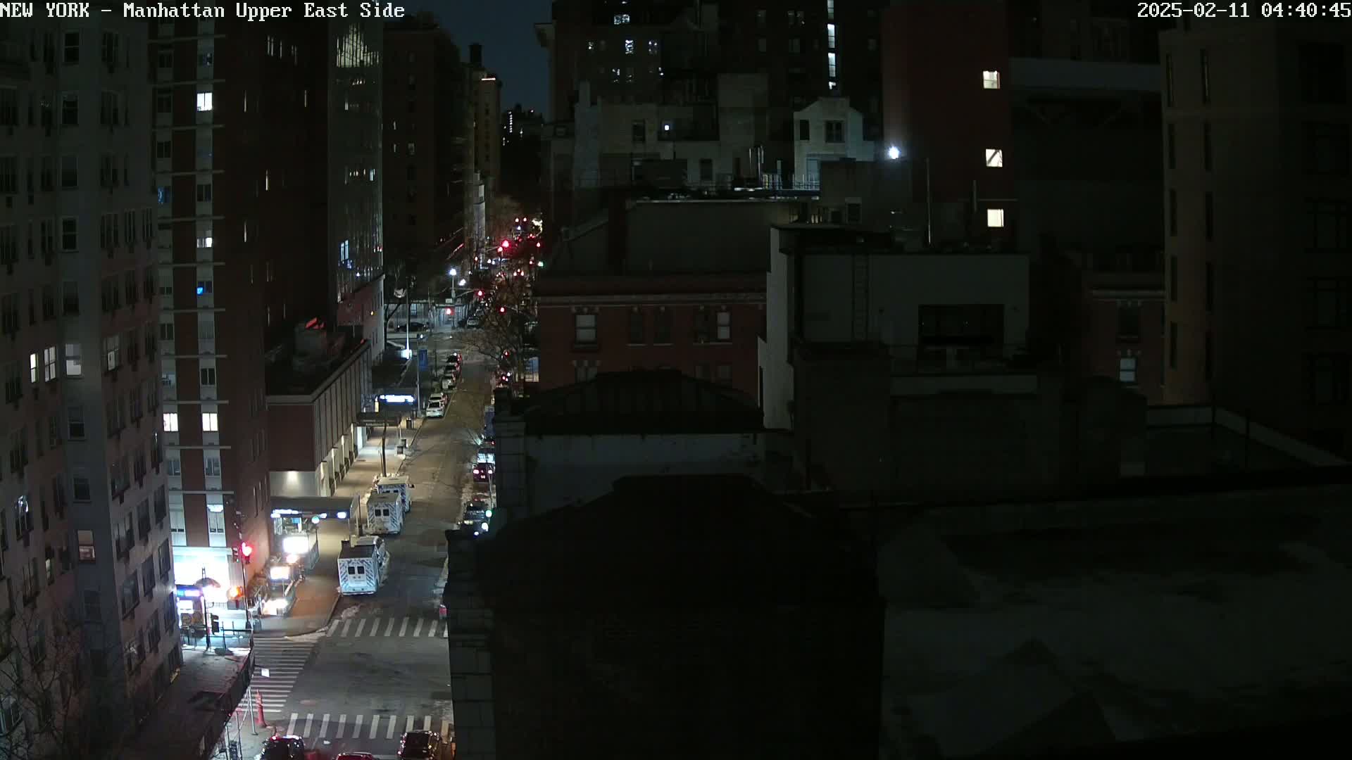 A nighttime, high-angle view of a city street with several buildings and sparse traffic under dark conditions.