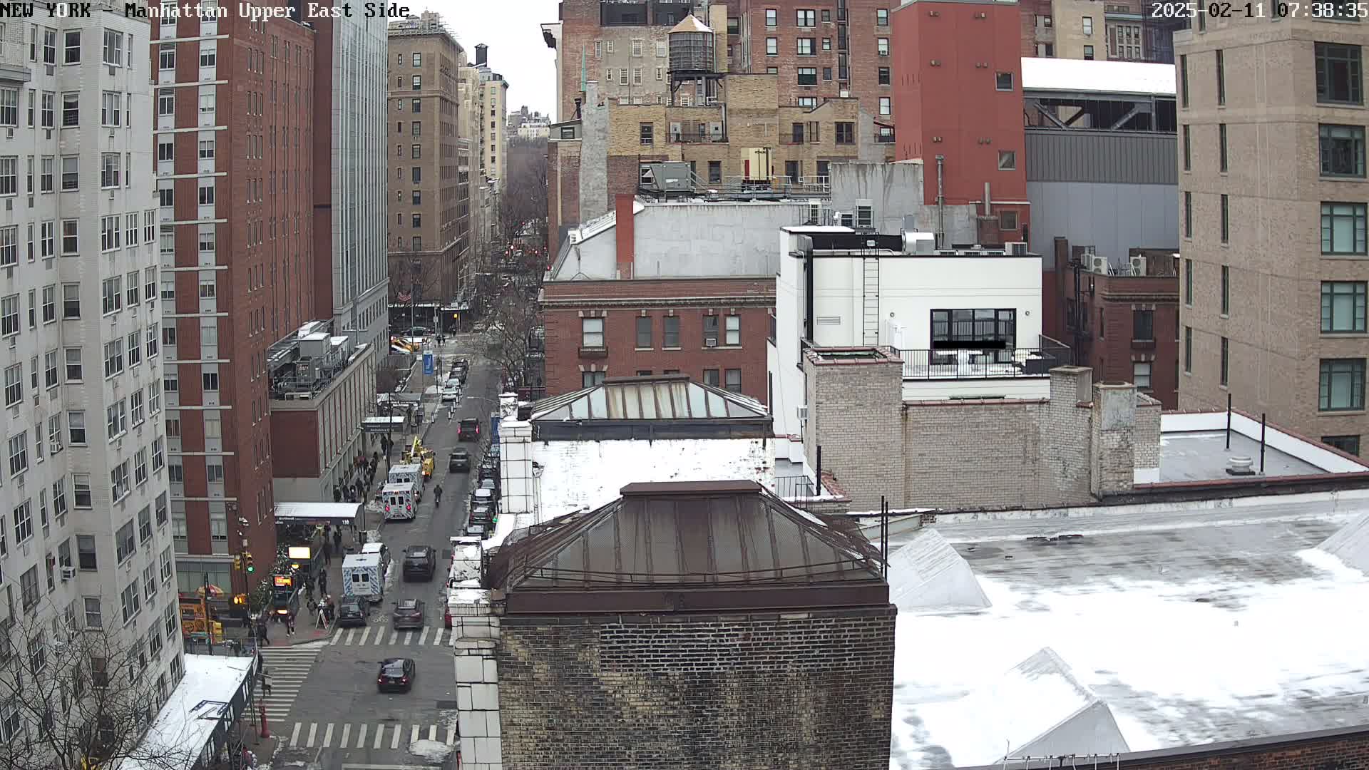 A snowy city street scene viewed from above shows several multi-story buildings and a street with cars and pedestrians.