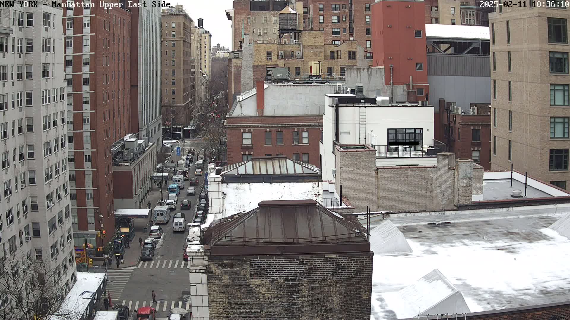 A snow-dusted cityscape view of a Manhattan street with multi-story buildings and traffic.