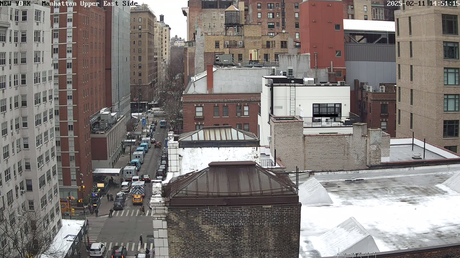 A snowy street scene in a dense city, showing several multi-story buildings and a street with some traffic.