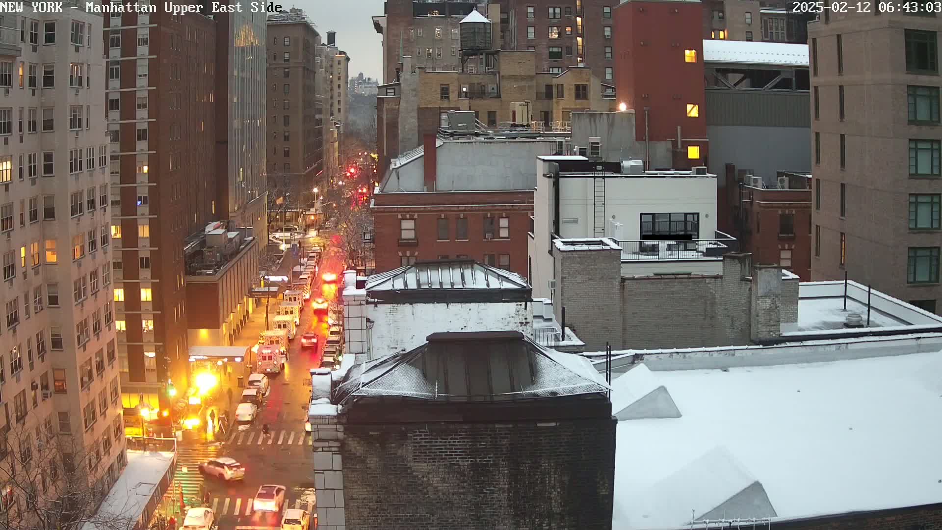 A snow-covered city street scene at twilight shows several city blocks with traffic and emergency vehicles.