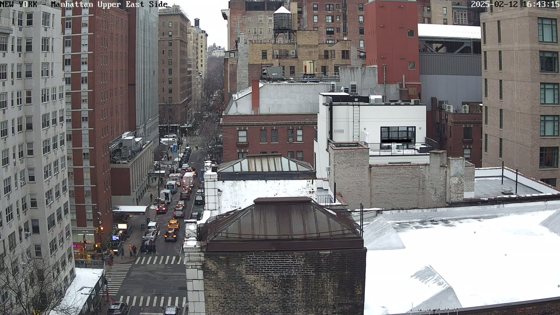 A snow-covered city street scene shows a view of several multi-story buildings, a busy street with cars, and some pedestrians at a crosswalk.